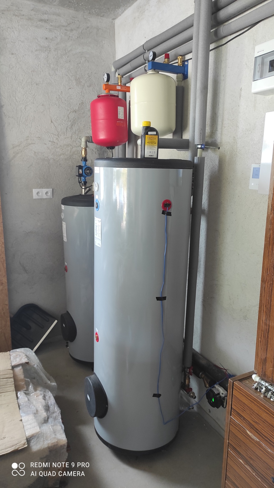 Dwa szare zasobniki ciepłej wody użytkowej w kotłowni, z widocznymi rurami w izolacji, zbiornikami wyrównawczymi i butelką preparatu MC1+ Protector na górze jednego z nich.