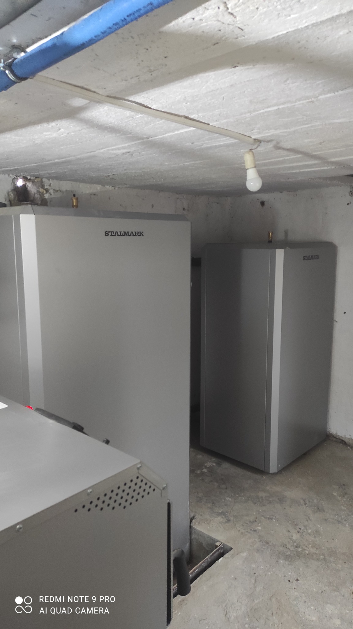 Dwie szare pompy ciepła Stalmark w piwnicy z betonową podłogą i sufitem, niebieska rura na suficie, surowa żarówka.