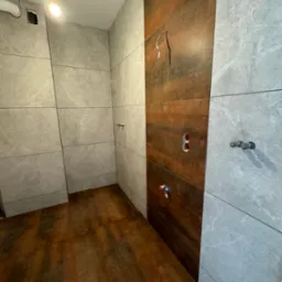 Łazienka w trakcie remontu, widoczne płytki imitujące marmur i drewno, przygotowane punkty instalacyjne, przew&oacute;d elektryczny wystający ze ściany, surowa żar&oacute;wka oświetlająca pomieszczenie.