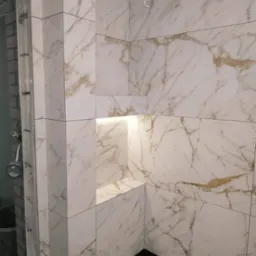 Wykończona wnęka z podświetleniem w kabinie prysznicowej, wyłożona białymi płytkami imitującymi marmur z delikatnym złotym użyleniem, widoczne czarne listwy podłogowe i fragment surowej ściany...