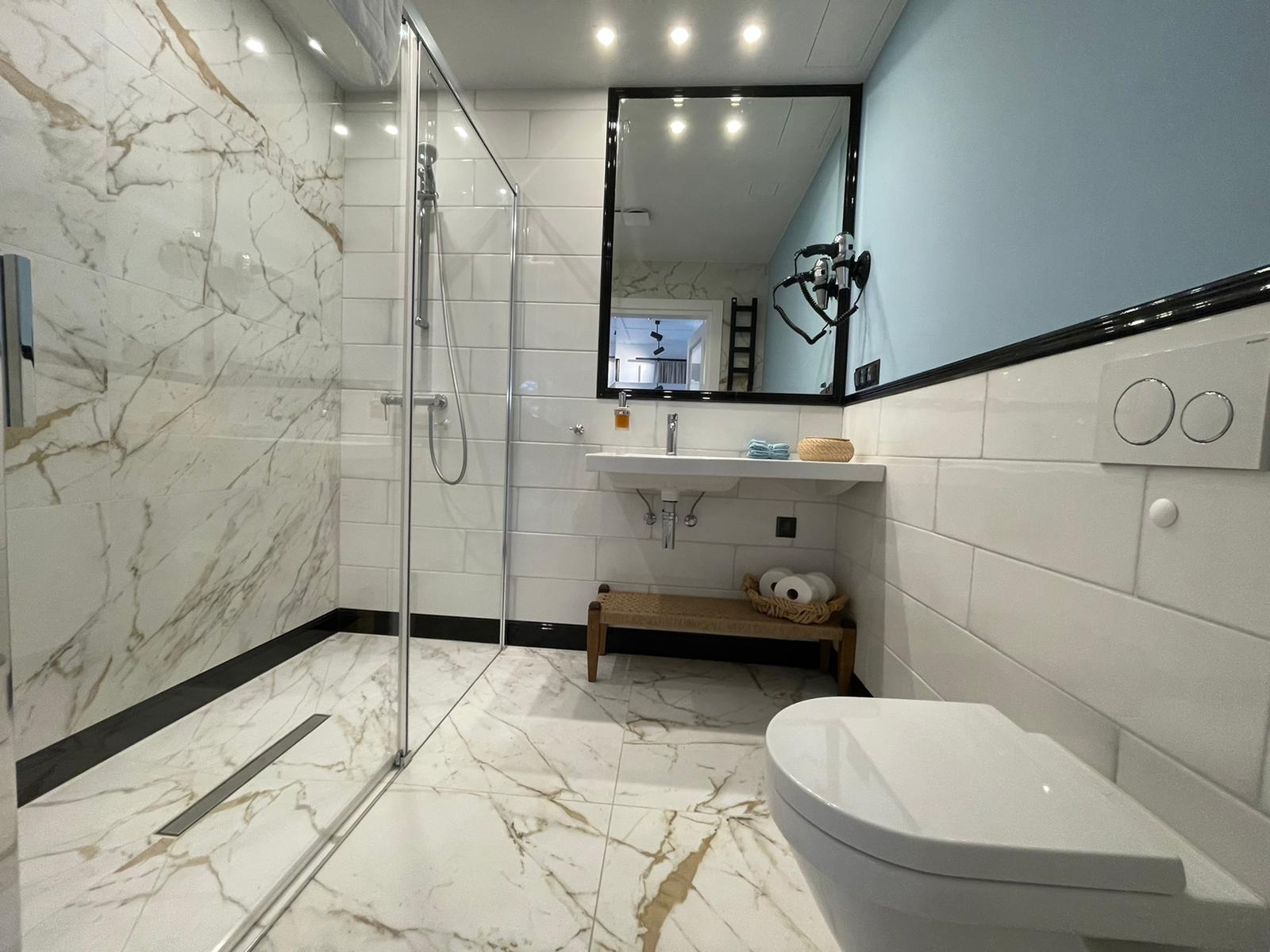 Nowoczesna łazienka z kabiną prysznicową typu walk-in, biało-złotymi płytkami imitującymi marmur na podłodze i ścianach, oraz białymi płytkami w cegiełkę. Umywalka z czarną ramą lustra, toaleta...