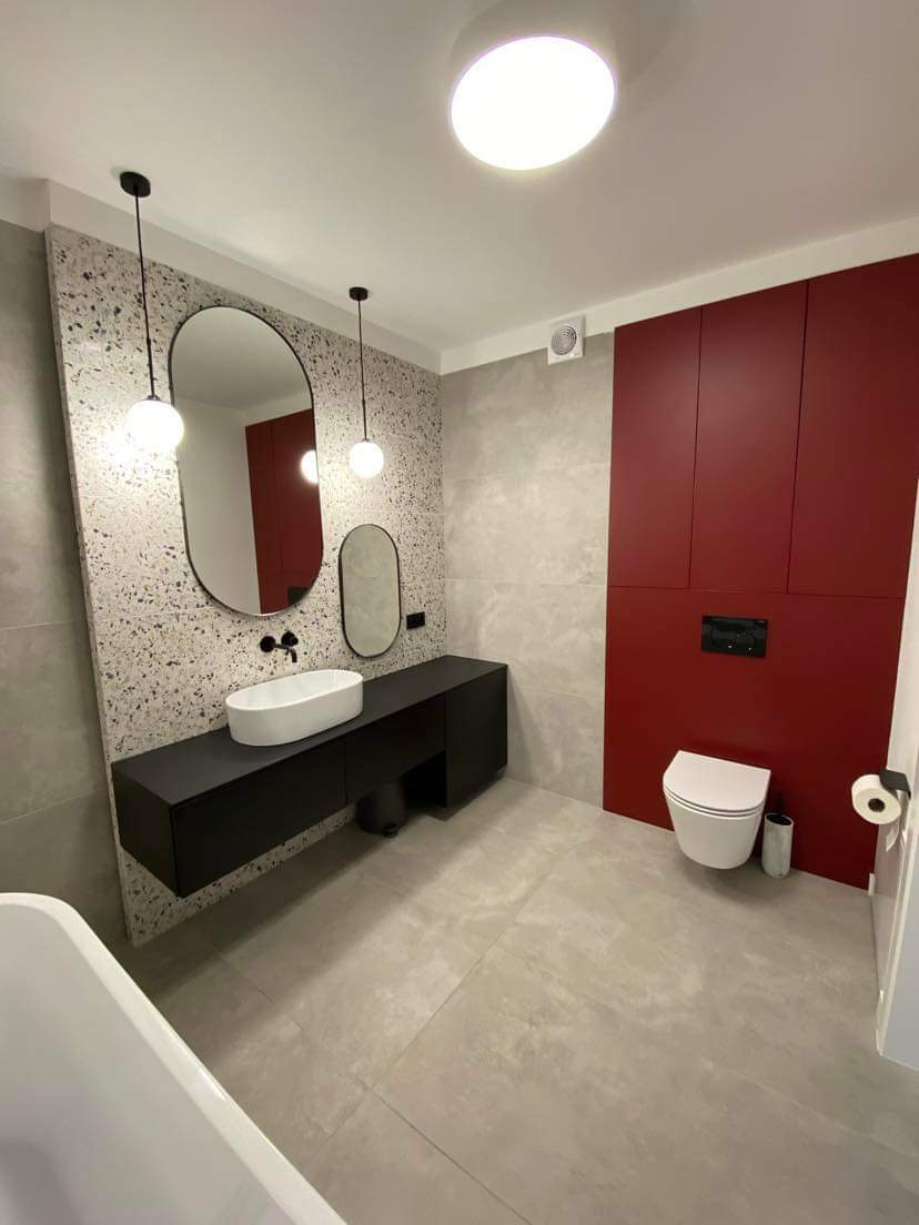 Nowoczesna łazienka z szarą terakotą na ścianie za umywalką, czarną szafką wiszącą z owalną białą umywalką, dwoma wiszącymi lampami kulami, dużym owalnym lustrem, czerwoną zabudową meblową...