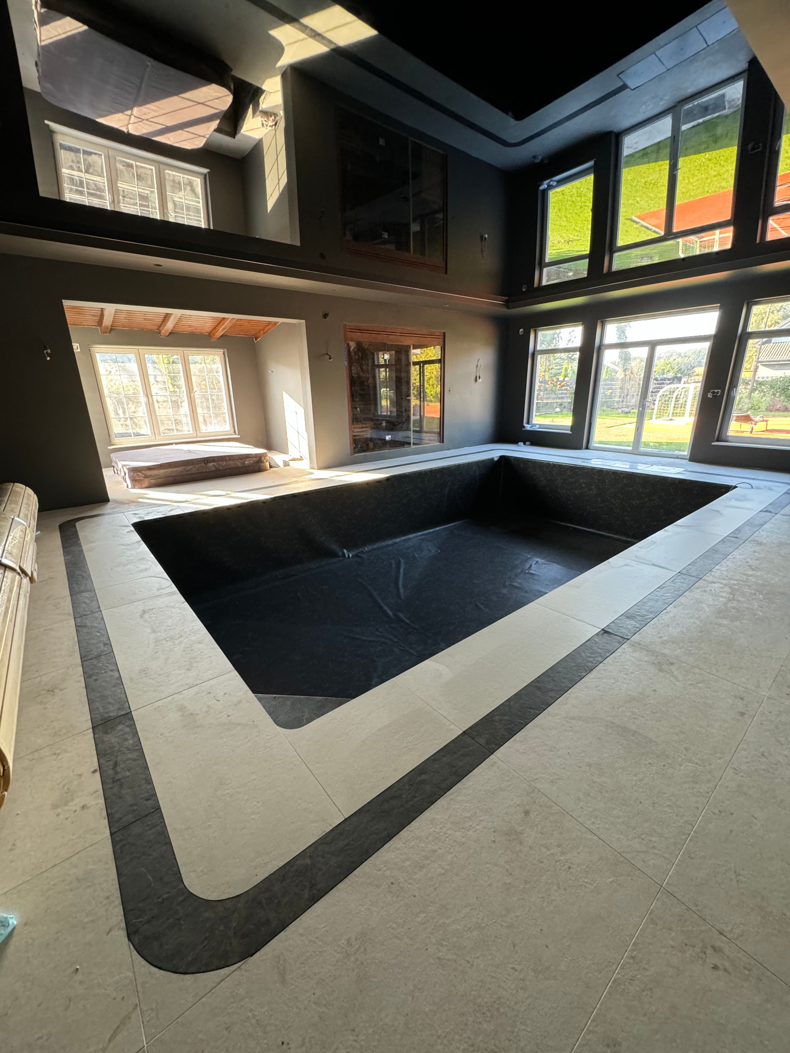Wnętrze w trakcie budowy z wyłożonymi płytkami podłogowymi i przygotowanym miejscem na basen z czarną folią wewnątrz, widoczne duże okna i surowe ściany.