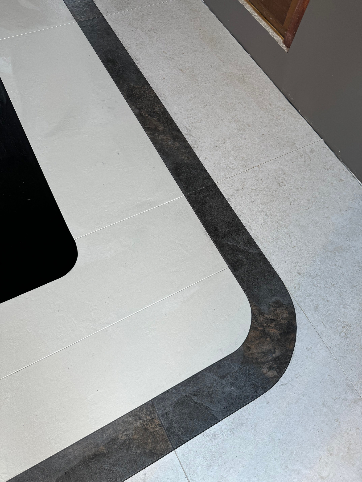 Wzorzysta podłoga z płytek w odcieniach bieli, szarości i czerni, z wyraźnym kontrastowym pasem w kształcie litery 'U' wykonanym z ciemnych płytek, tworzącym dekoracyjny element na jasnej powierzchni.