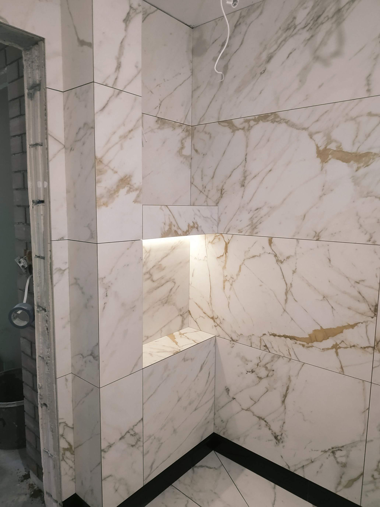 Wykończona wnęka z podświetleniem w kabinie prysznicowej, wyłożona białymi płytkami imitującymi marmur z delikatnym złotym użyleniem, widoczne czarne listwy podłogowe i fragment surowej ściany...