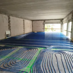 Niebieskie rury ogrzewania podłogowego ułożone w pętle na podkładzie z siatką w surowym, niewykończonym pomieszczeniu z widocznymi pustakami i betonowym stropem.