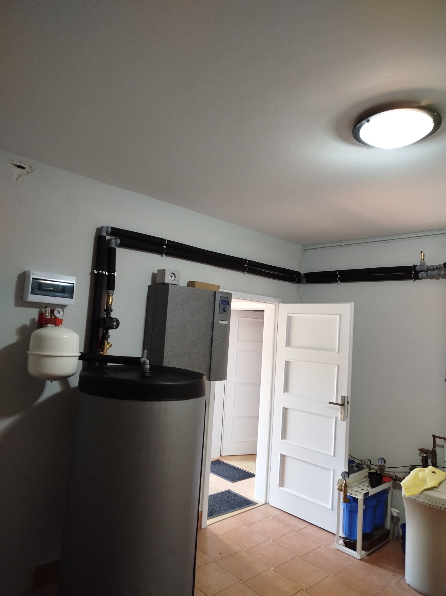 Wnętrze kotłowni z szarym zasobnikiem ciepłej wody, czarnymi rurami CO biegnącymi pod sufitem, skrzynką elektryczną i filtrami wody przy otwartych, białych drzwiach.