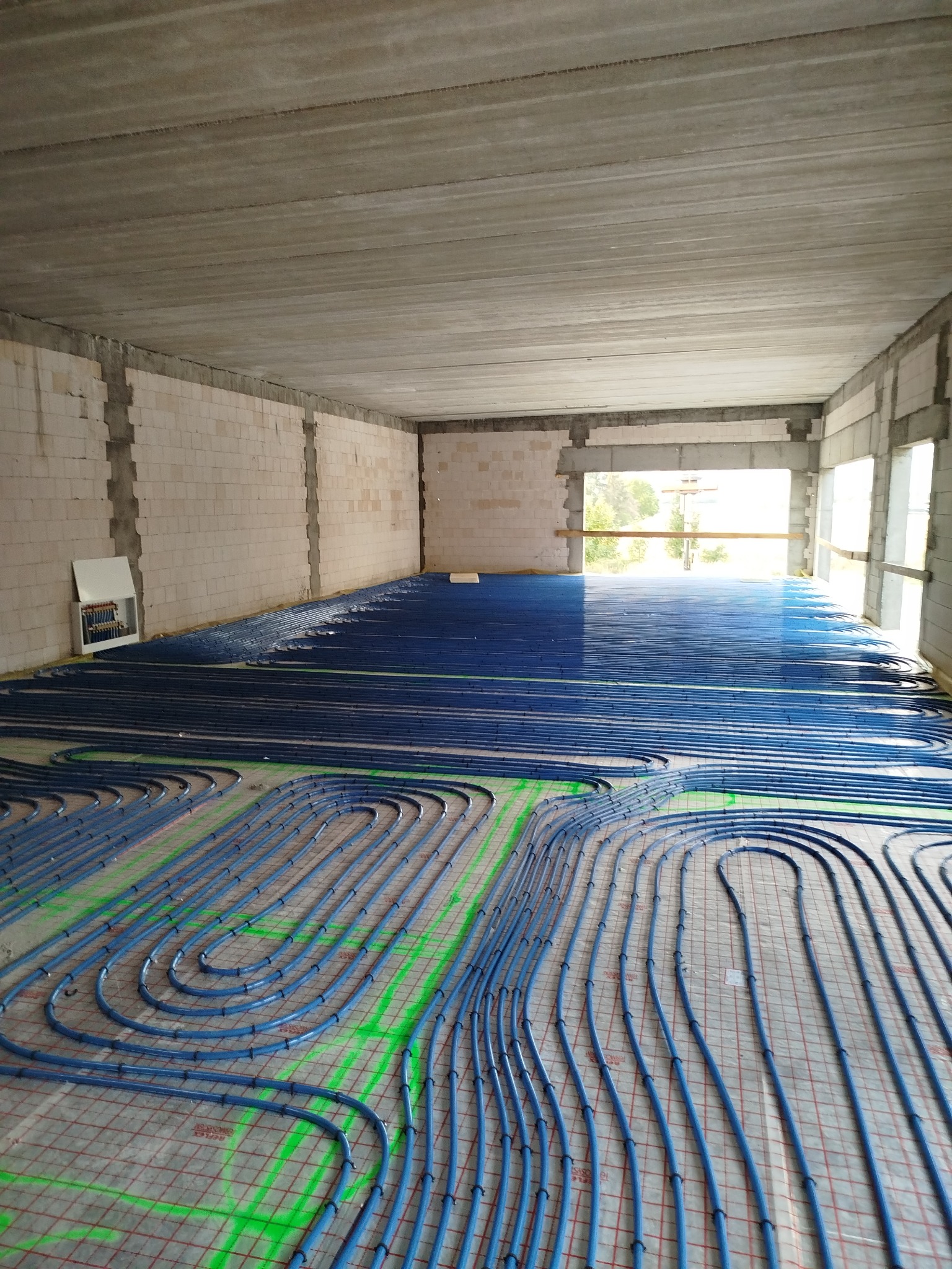 Niebieskie rury ogrzewania podłogowego ułożone w pętle na podkładzie z siatką w surowym, niewykończonym pomieszczeniu z widocznymi pustakami i betonowym stropem.