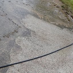 Michał Chojnacki - Fragment czyszczonej, mokrej kostki brukowej z widocznym wężem. Częściowo umyta powierzchnia kontrastuje z zabrudzoną. Widoczne ślady ziemi i trawy.