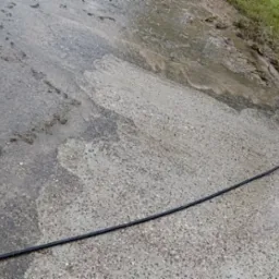 Fragment czyszczonej, mokrej kostki brukowej z widocznym wężem. Częściowo umyta powierzchnia kontrastuje z zabrudzoną. Widoczne ślady ziemi i trawy.