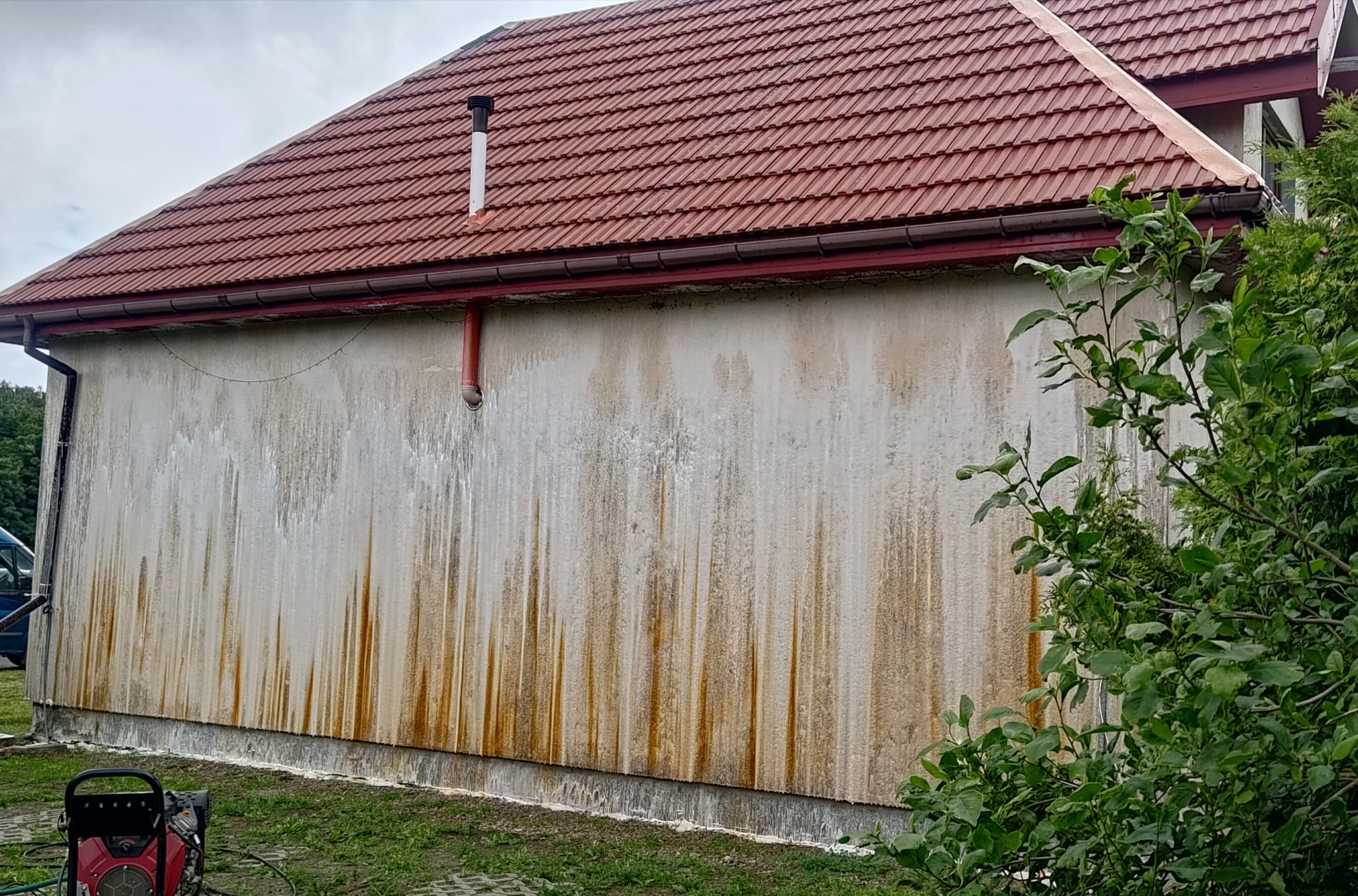 Zabrudzona elewacja budynku z czerwonym dachem, widoczne zacieki i plamy. W tle zieleń, w dolnym rogu myjka ciśnieniowa. Ujęcie z bliska, przed czyszczeniem.
