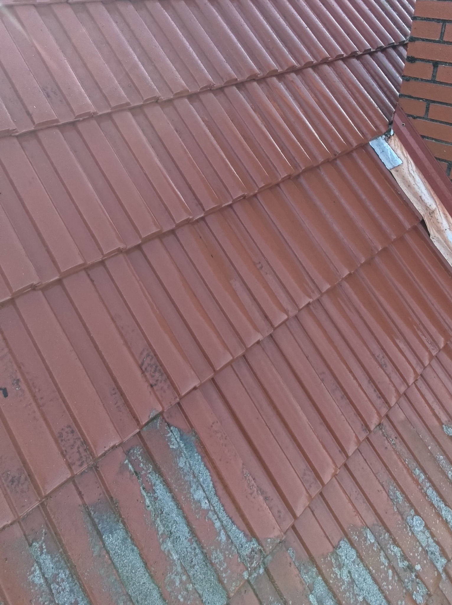 Czerwona dachówka ceramiczna, częściowo pokryta wilgocią i mchem, widoczny fragment komina z cegły w prawym górnym rogu. Ujęcie z góry.