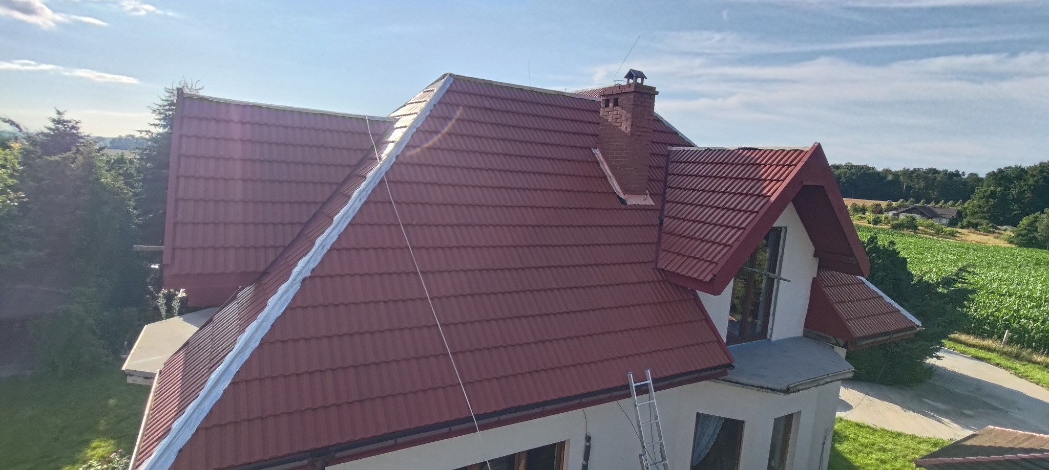 Czerwony dach z dachówki karpiówki na dwukondygnacyjnym domu, widok z góry. Na dachu komin z cegły i aluminiowa drabina oparta o ścianę. W tle pole uprawne i drzewa.