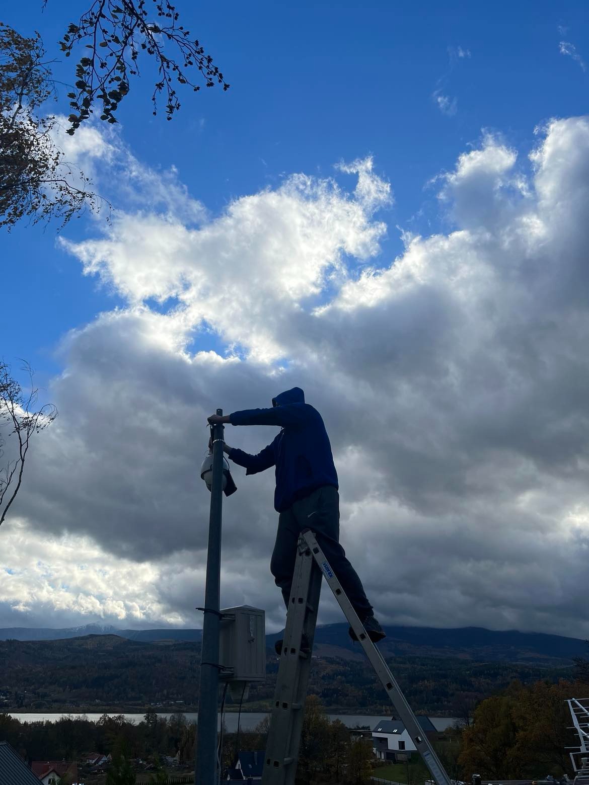 Instalator na drabinie montuje kamerę monitoringu na słupie, w tle widok na góry i jezioro pod pochmurnym niebem.