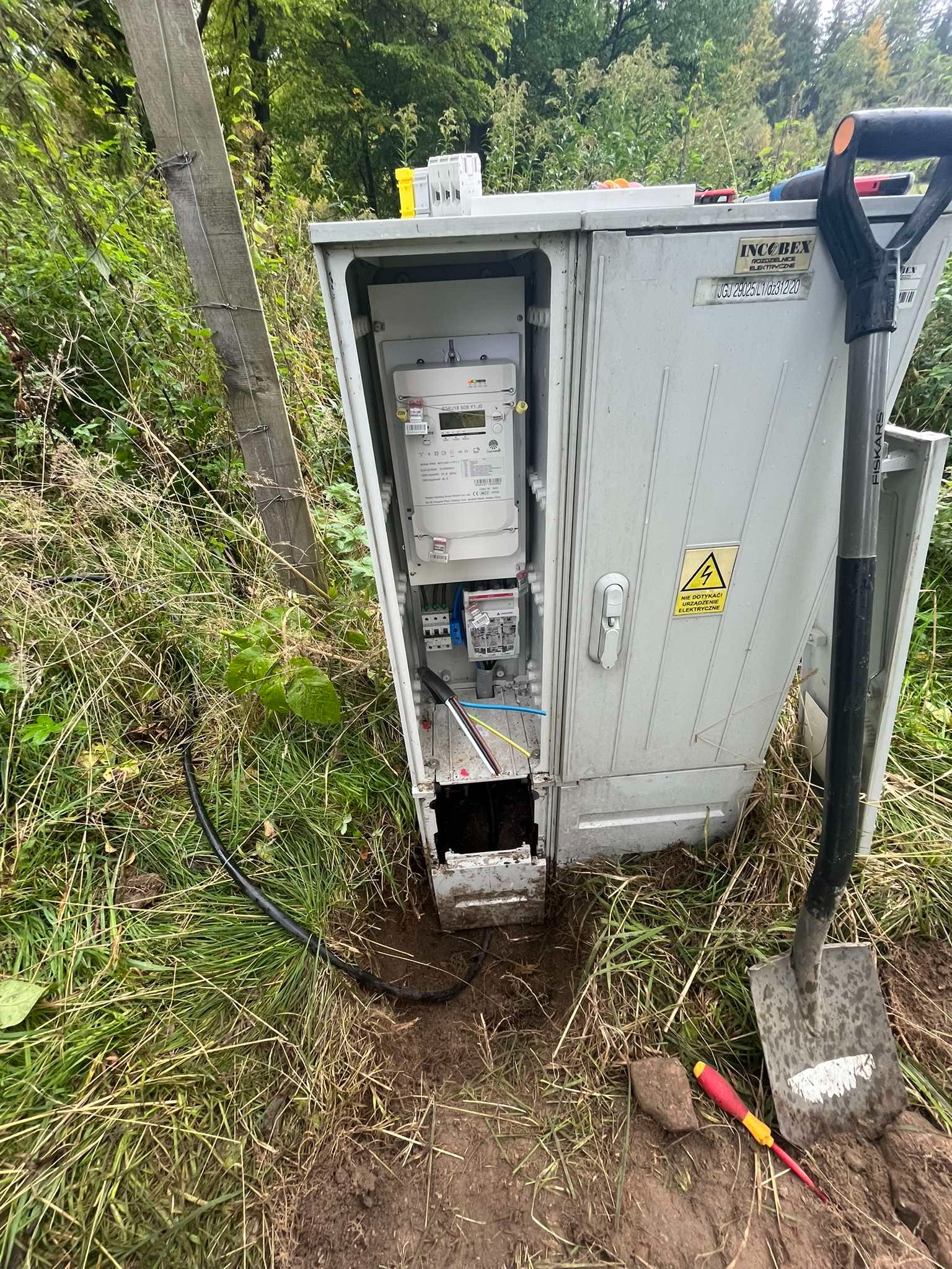 Otwarta skrzynka elektryczna z licznikiem, wystająca z ziemi na zewnątrz, obok leży szpadel i śrubokręt, widoczne kable i ostrzeżenie o wysokim napięciu.