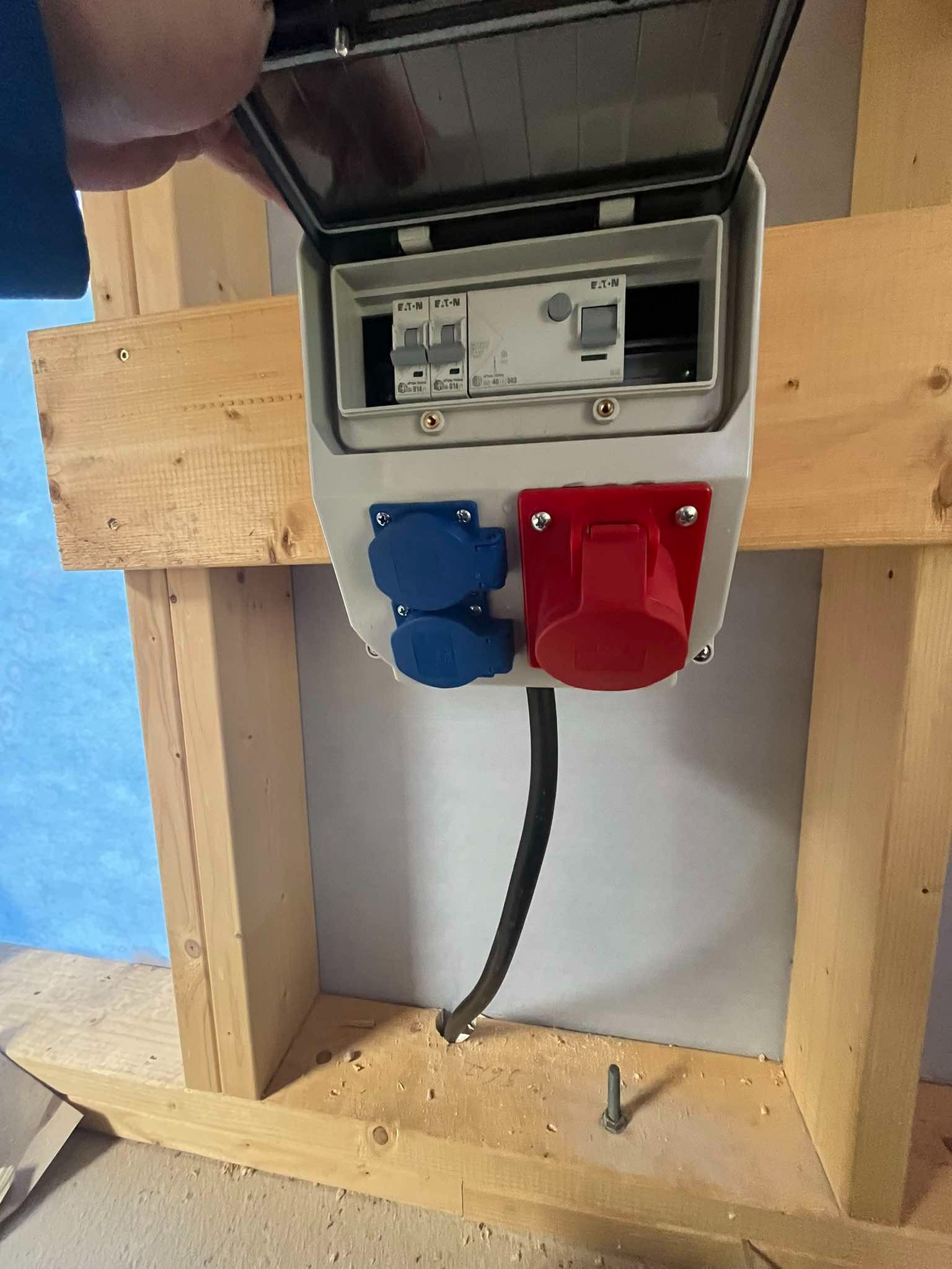 Otwarta skrzynka elektryczna z bezpiecznikami EATON i gniazdami siłowymi (czerwone i niebieskie) zamontowana na drewnianej konstrukcji szkieletowej ściany.
