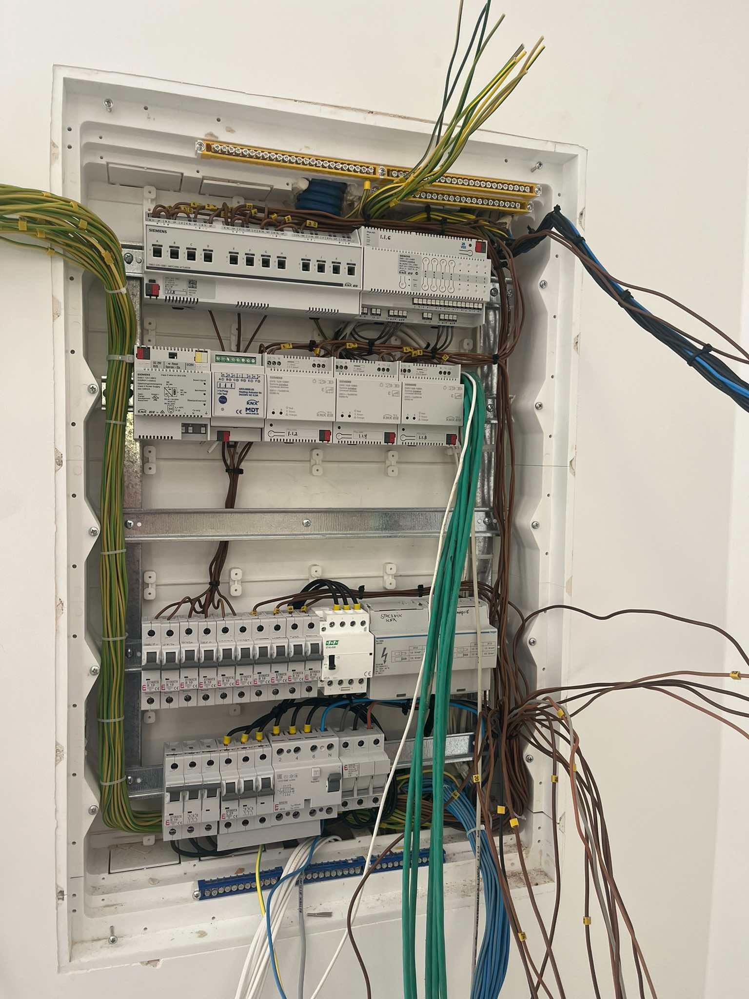 Otwarta skrzynka elektryczna z widocznymi bezpiecznikami, przekaźnikami, przewodami o różnych kolorach i oznaczeniach, zamontowana na białej ścianie. Część przewodów jest uporządkowana, a część...