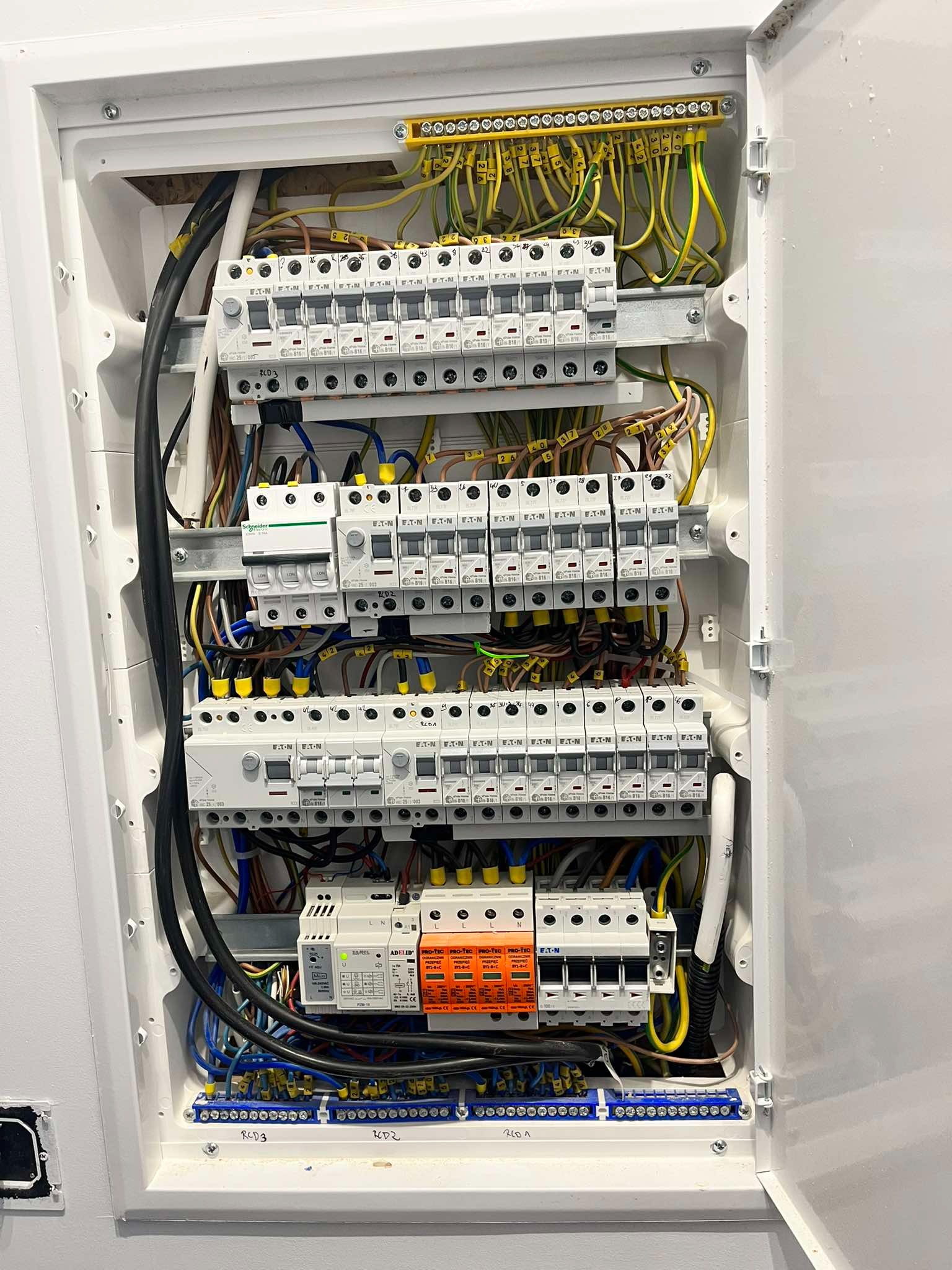 Otwarta skrzynka elektryczna z bezpiecznikami, przewodami oznaczonymi numerami i listwami zaciskowymi w kolorach żółto-zielonym i niebieskim, zamontowana na białej ścianie.