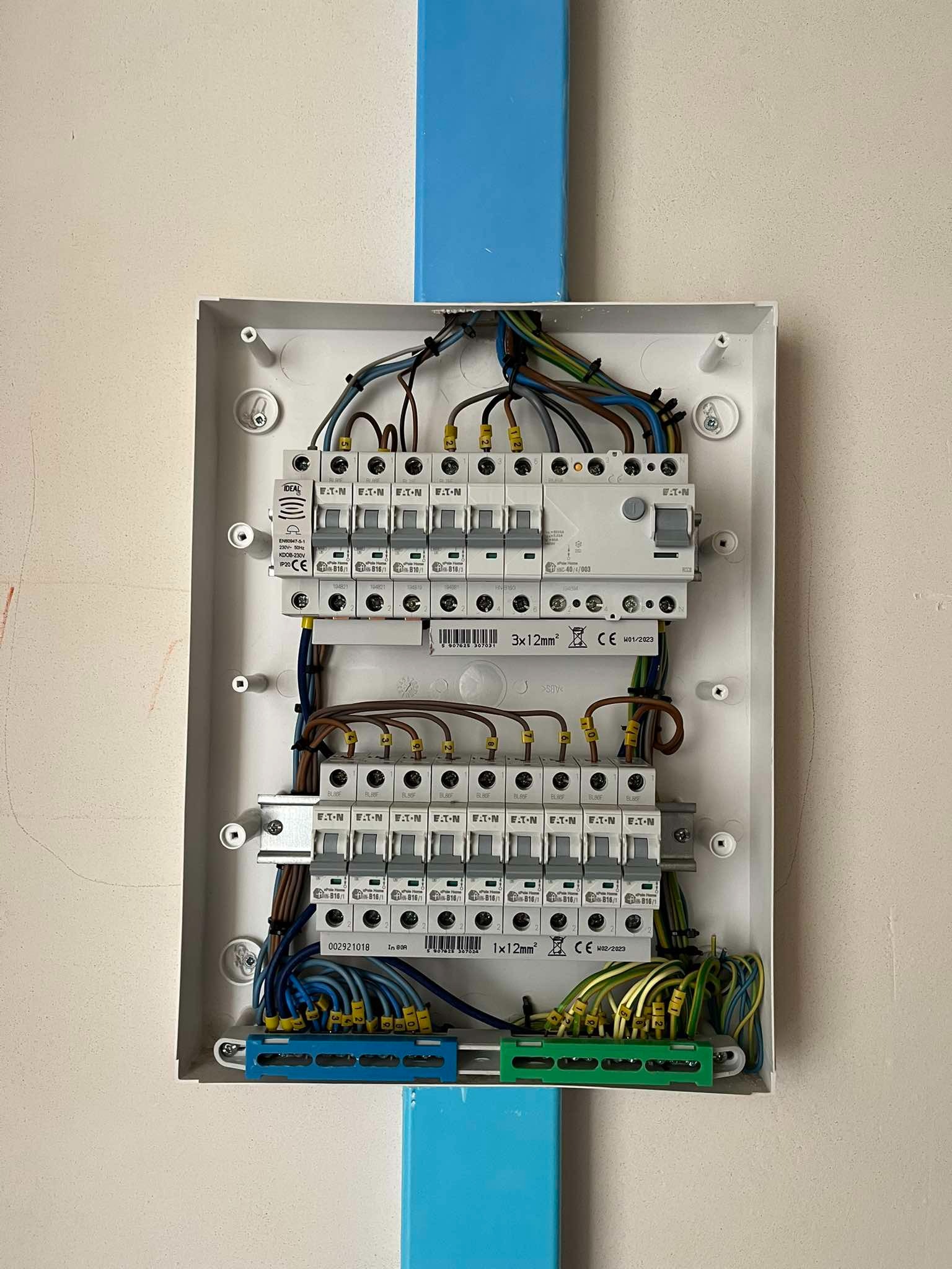 Otwarta skrzynka bezpiecznikowa Eaton z podłączonymi przewodami o różnych kolorach, widoczne oznaczenia na przewodach i listwach zaciskowych, zamontowana na ścianie z niebieskim pionowym elementem...