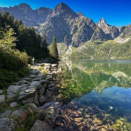 Szymon Piechowiak - Malowniczy krajobraz górski z jeziorem i ścieżką w Bydgoszczy, woda jeziora odbija szczyty górskie, na ścieżce w oddali widoczna sylwetka człowieka.