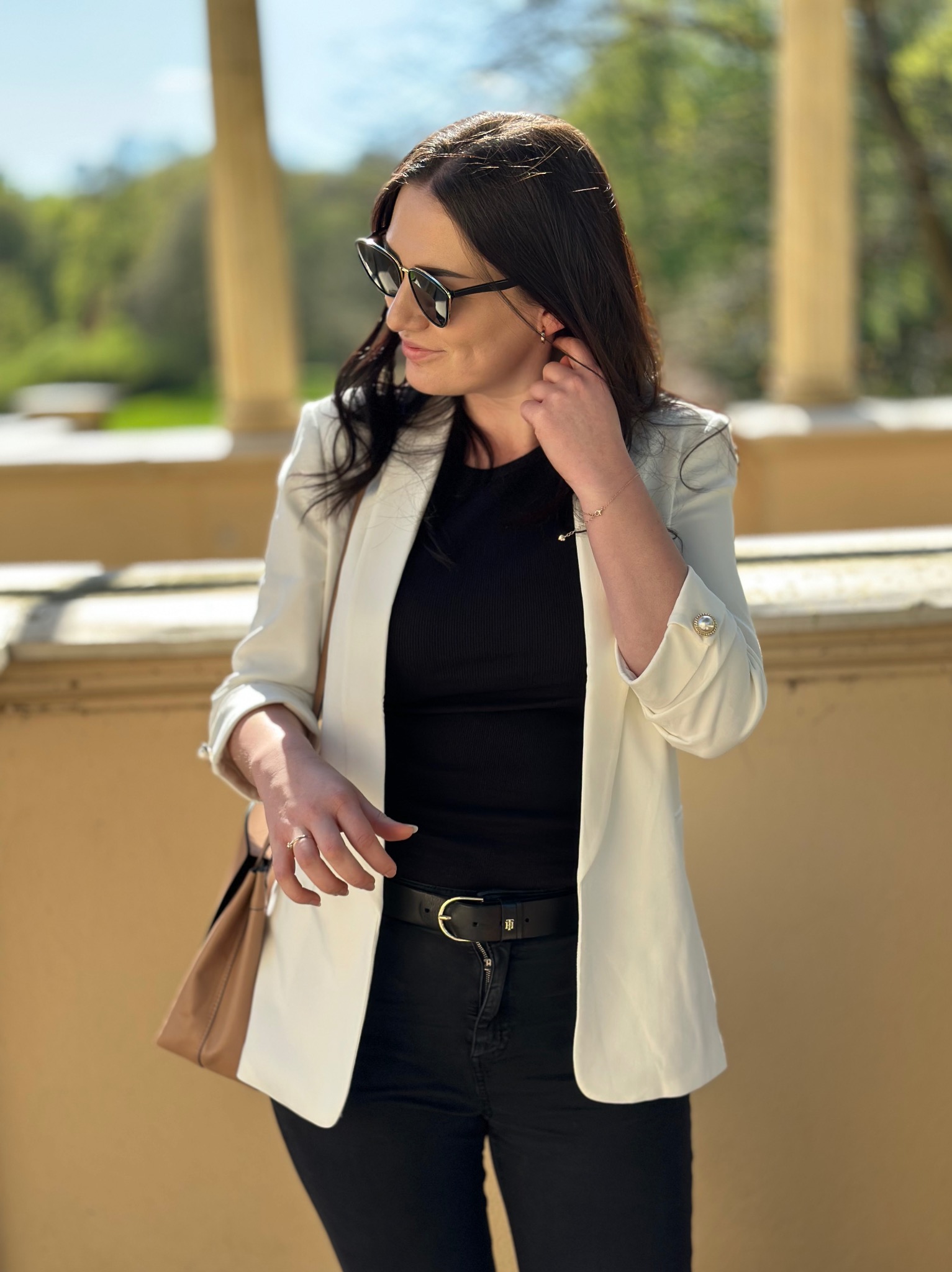 Kobieta w okularach przeciwsłonecznych, białej marynarce i czarnym topie pozuje na tle kolumn i zieleni w słoneczny dzień.