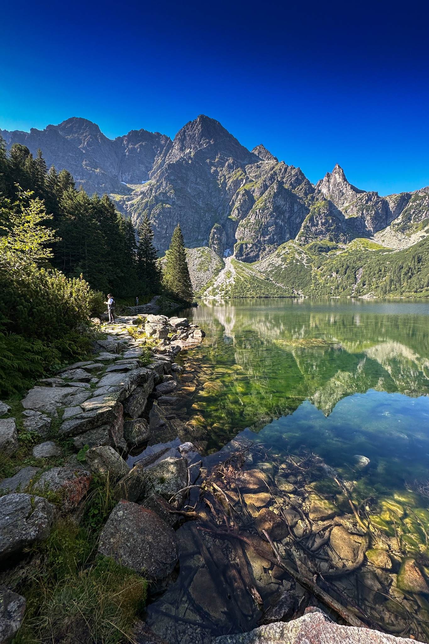 Malowniczy krajobraz górski z jeziorem i ścieżką w Bydgoszczy, woda jeziora odbija szczyty górskie, na ścieżce w oddali widoczna sylwetka człowieka.