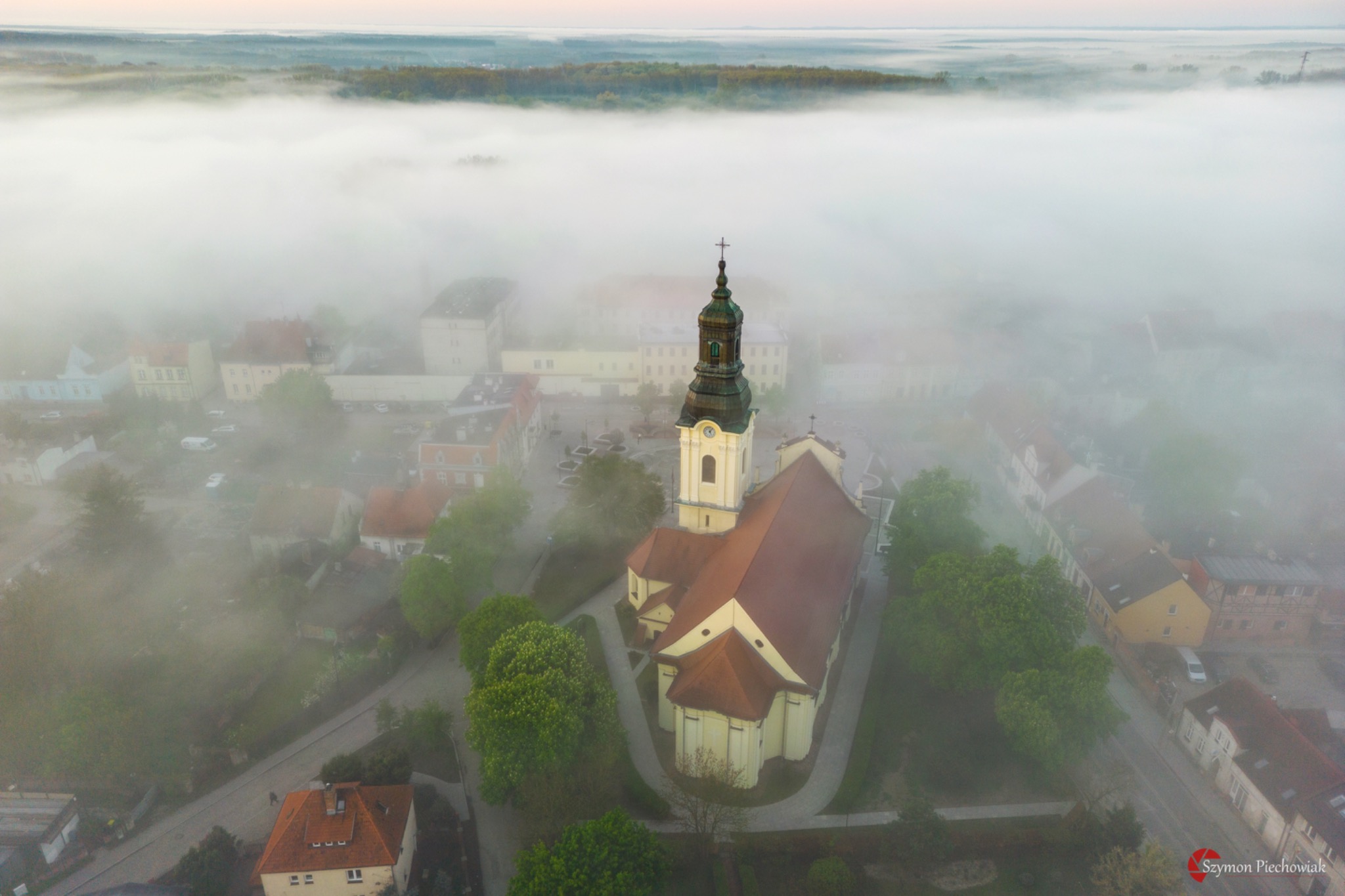 Widok z drona na kościół z wieżą i zegarem, spowity poranną mgłą, z widocznymi budynkami miasta i lasem w tle.
