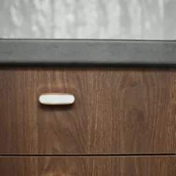 Szczegółowe ujęcie ciemnobrązowej drewnianej szafki z dwoma szufladami i marmurowymi uchwytami w złotej oprawie, na blacie z szarego kamienia, z rozmytym tłem w odcieniach szarości.