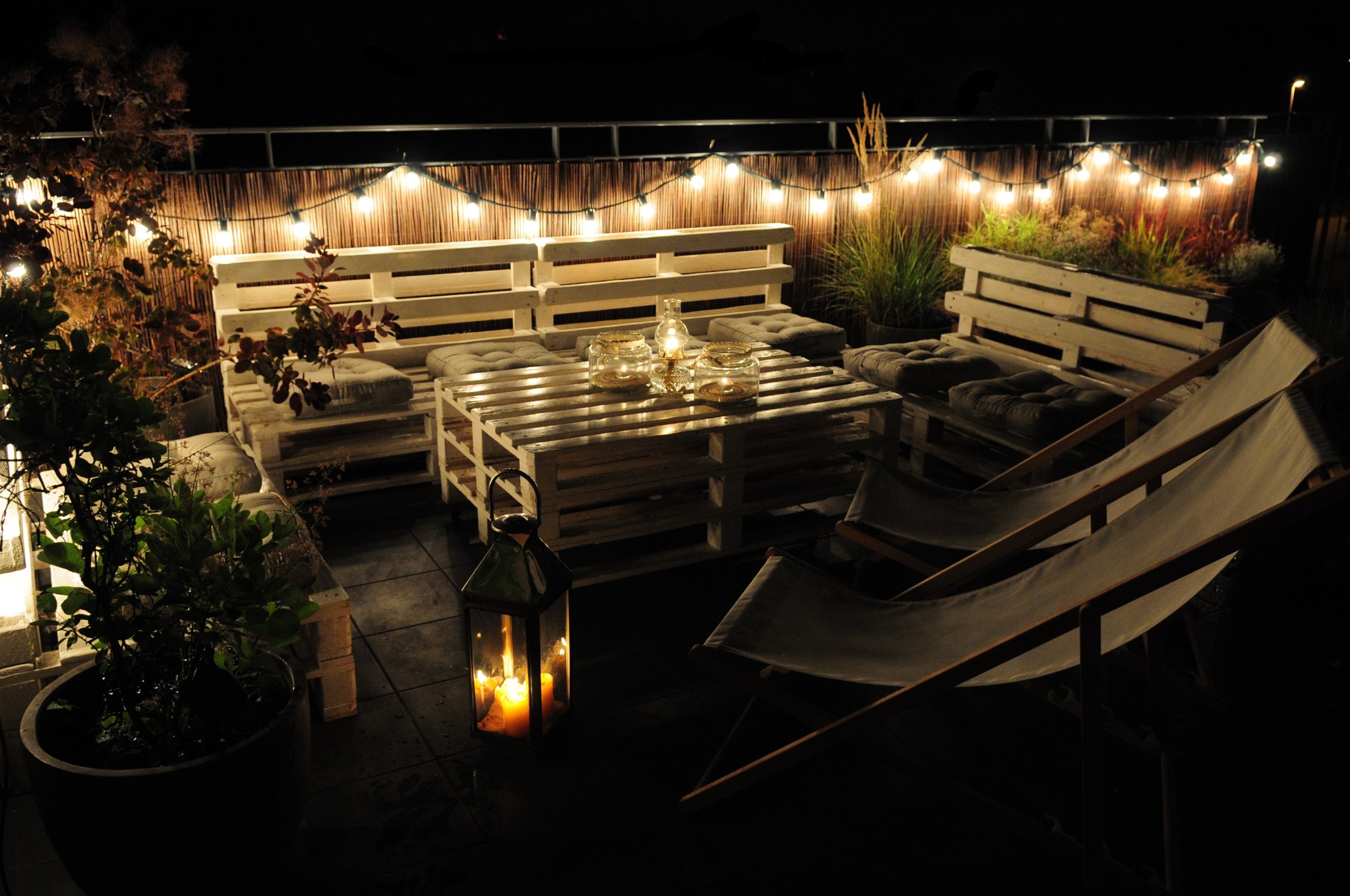 Oświetlony taras nocą z meblami z palet, lampionem ze świecami i leżakami, tworzący strefę wypoczynkową i jadalnianą.