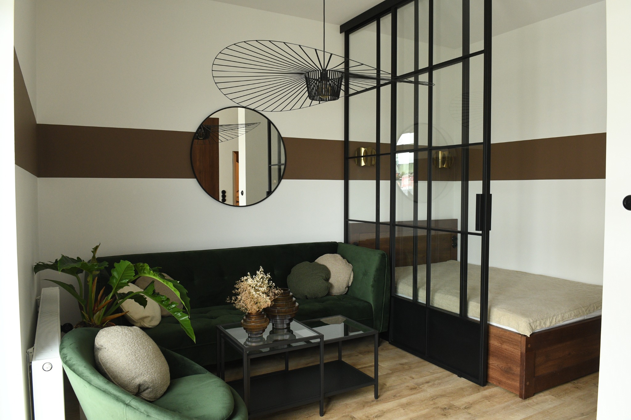 Nowoczesny salon z zieloną sofą, stolikiem kawowym z dekoracjami florystycznymi, okrągłym lustrem na ścianie z brązowym pasem i przeszkloną ścianą oddzielającą sypialnię z łóżkiem.