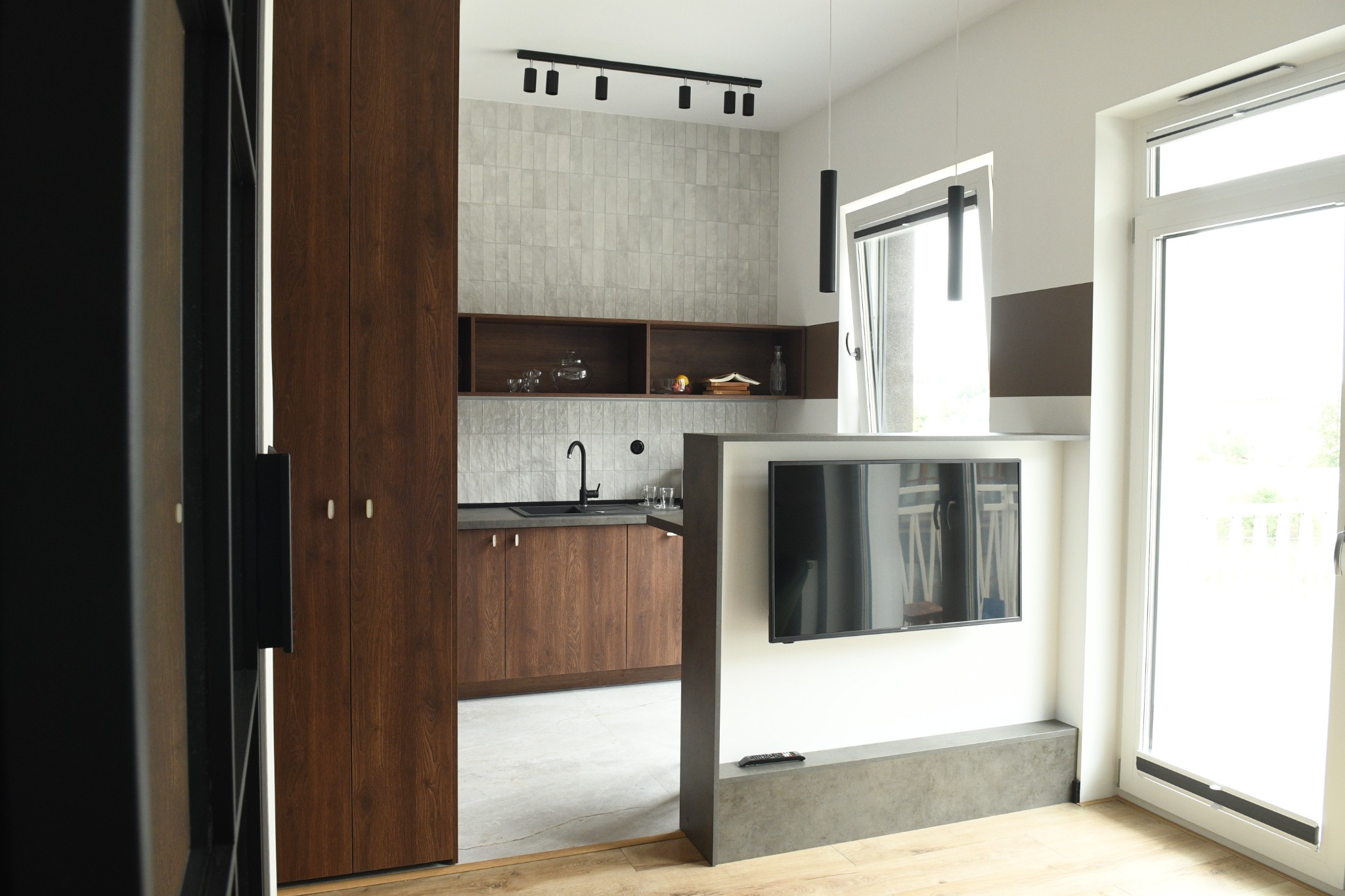 Nowoczesna kuchnia z drewnianymi szafkami, szarą ścianą z płytek i telewizorem na ścianie, widok z perspektywy wejścia, jasne wnętrze z dużym oknem balkonowym.