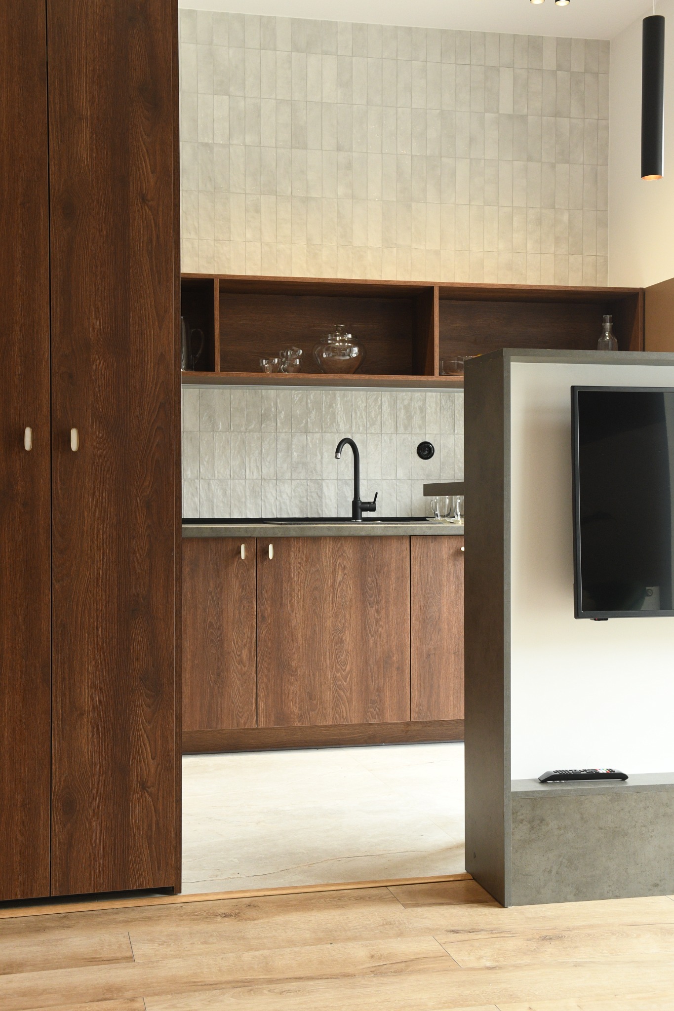Nowoczesna kuchnia z drewnianymi szafkami, czarną armaturą i dekoracyjnymi płytkami ściennymi, widok z salonu z telewizorem i pilotem leżącym na półce.