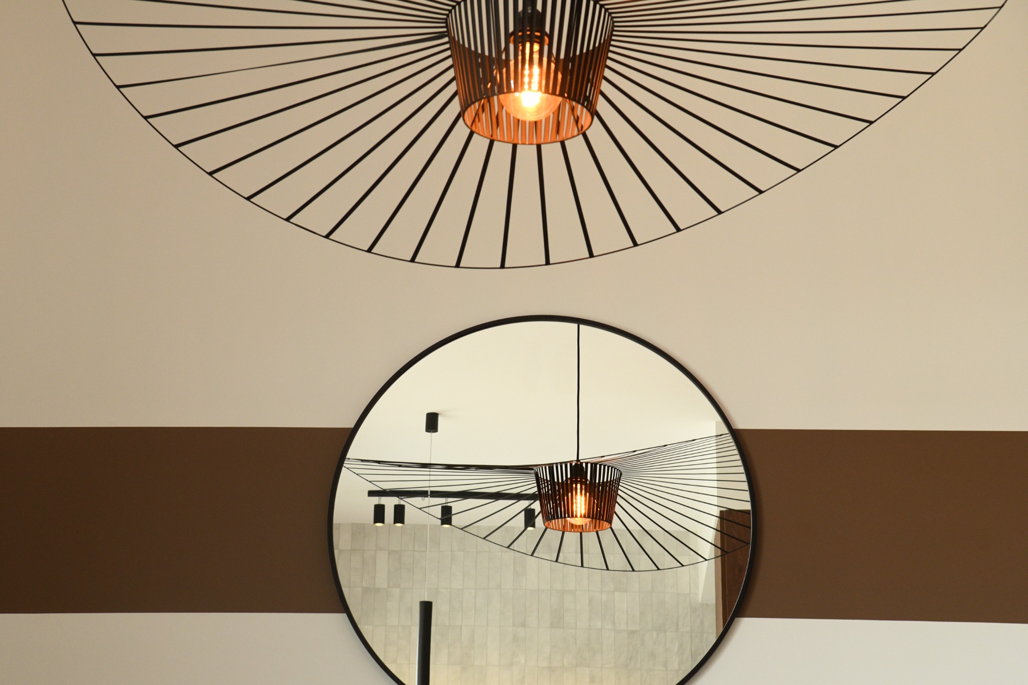 Nowoczesna lampa wisząca z metalowym kloszem i okrągłe lustro w czarnej ramie na ścianie z poziomym pasem w kolorze brązowym.