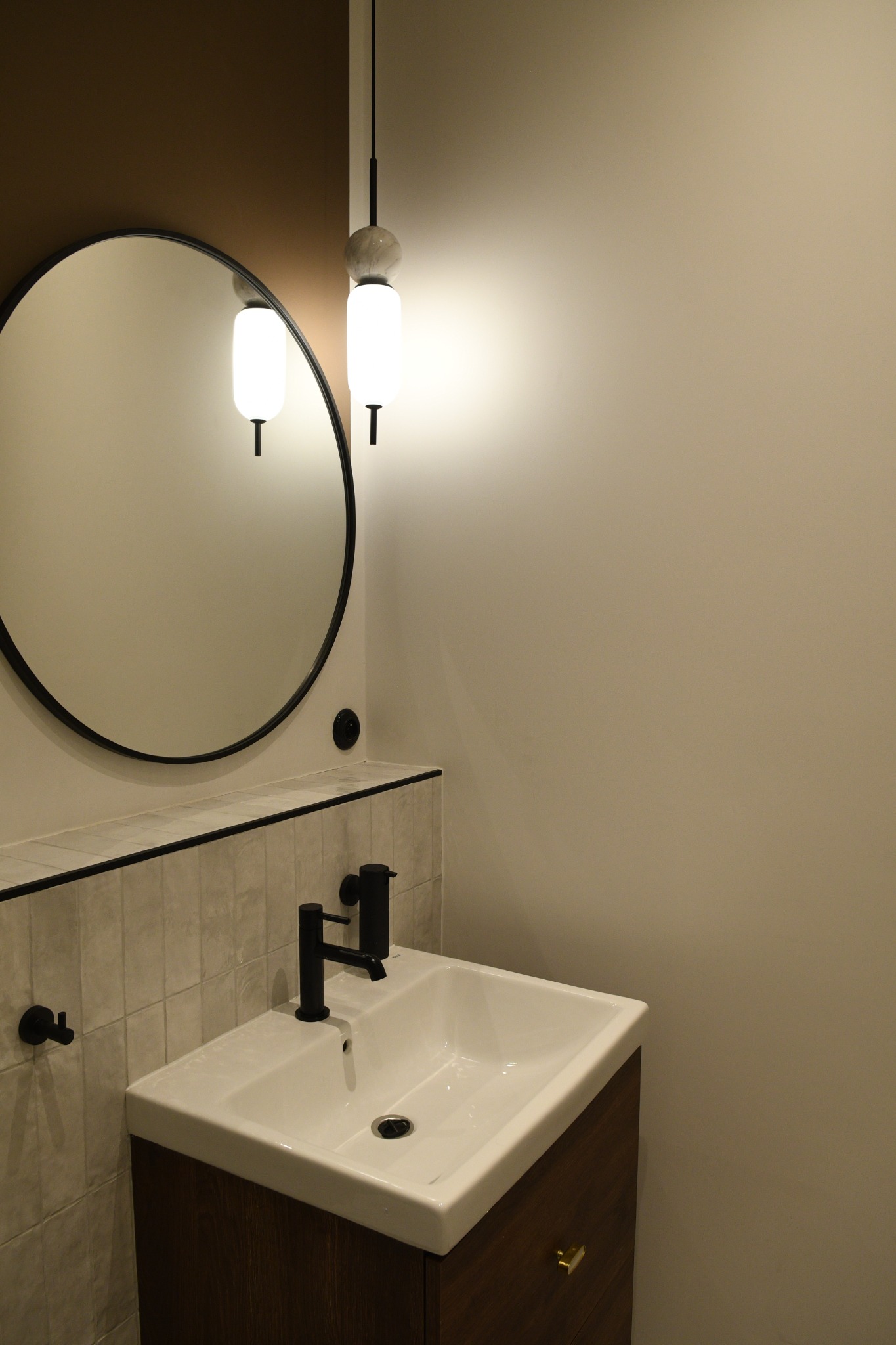 Nowoczesna łazienka z umywalką nablatową, czarną armaturą, okrągłym lustrem w czarnej ramie i wiszącą lampą z mlecznego szkła.