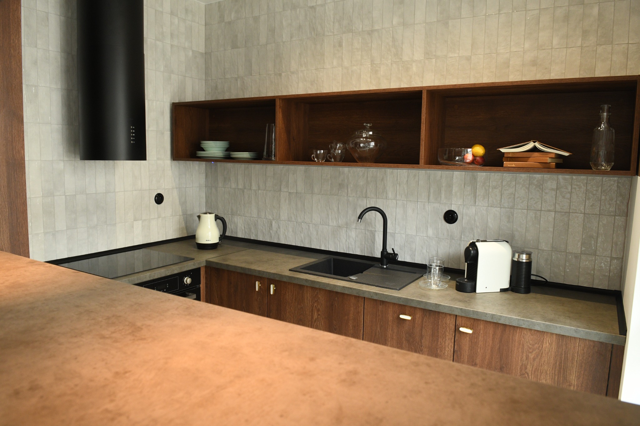 Nowoczesna kuchnia z drewnianymi szafkami, szarymi płytkami na ścianie i otwartymi półkami z dekoracjami, czarny okap nad płytą indukcyjną, ekspres do kawy na blacie.