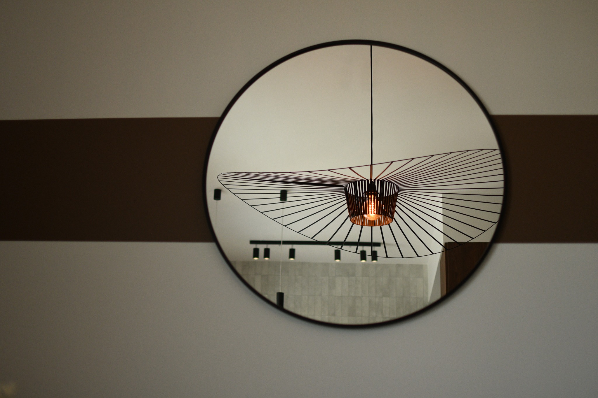 Okrągłe lustro z czarną ramą na ścianie z dwukolorowym pasem, odbijające nowoczesną lampę wiszącą z metalowymi prętami i żarówką oraz fragment sufitu z oświetleniem punktowym.