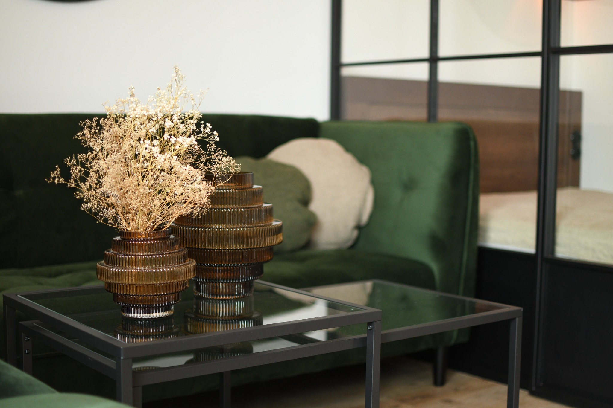 Dwie dekoracyjne, prążkowane, brązowe wazy na szklanym stoliku kawowym, z suszonymi kwiatami, na tle zielonej, aksamitnej sofy i szklanej ściany działowej.