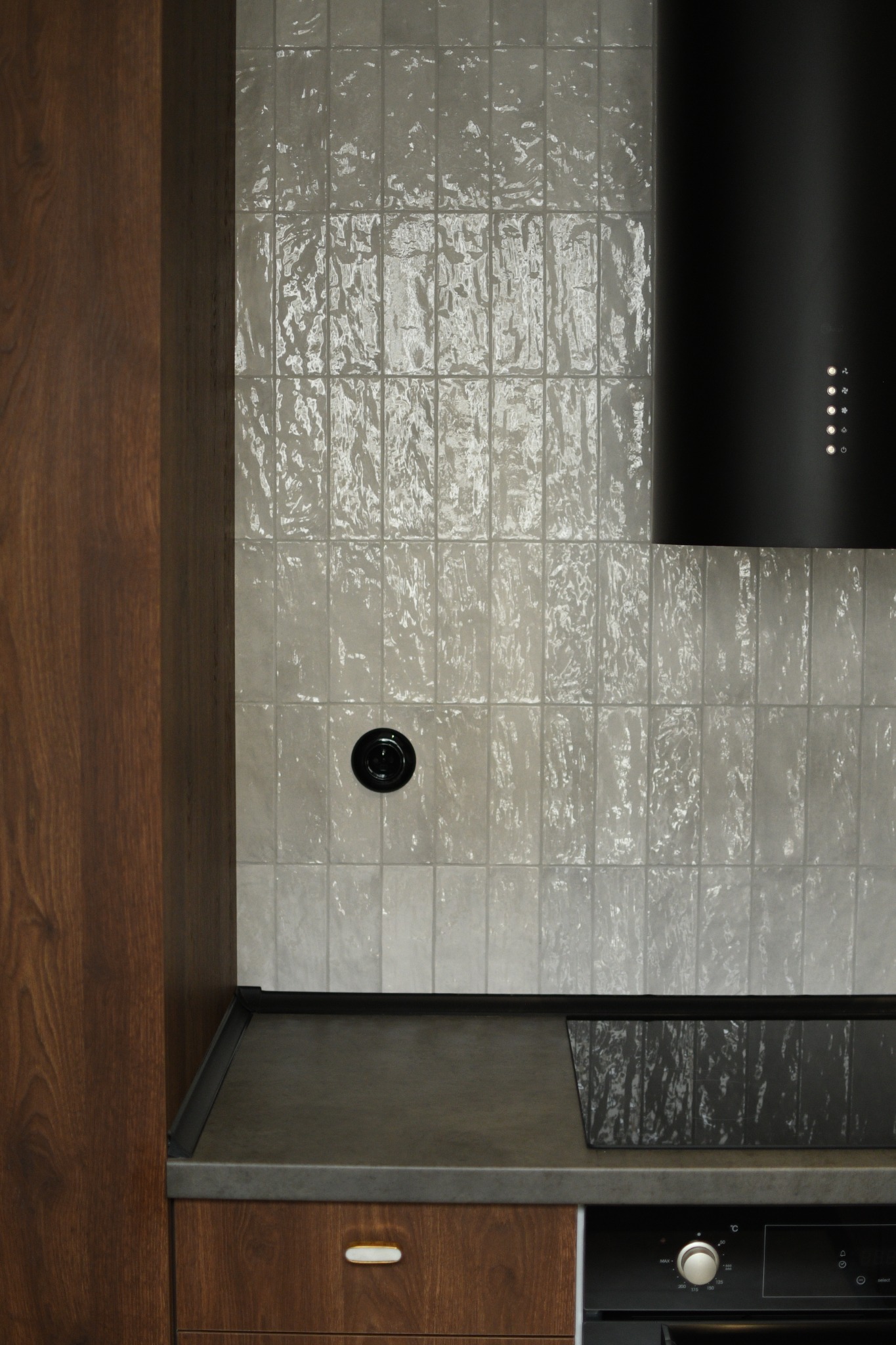 Fragment kuchni z drewnianymi szafkami, szarym blatem, ceramicznymi płytkami o nieregularnej fakturze i czarnym okapem z oświetleniem LED.