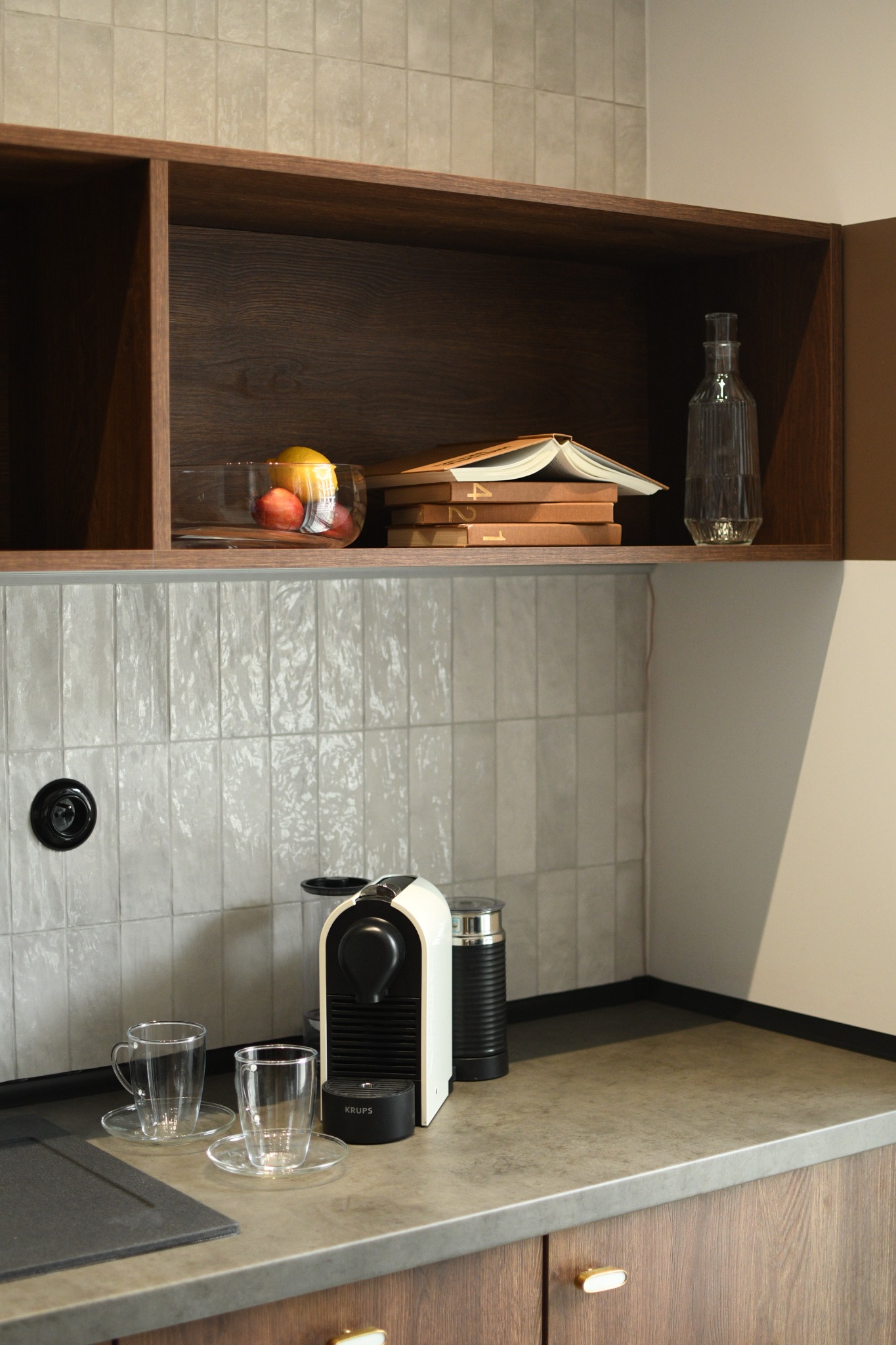Nowoczesna kuchnia z drewnianymi szafkami, szarym blatem i białymi płytkami na ścianie, ekspres do kawy Krups, szklanki, owoce w szklanej misce i książki na półce.