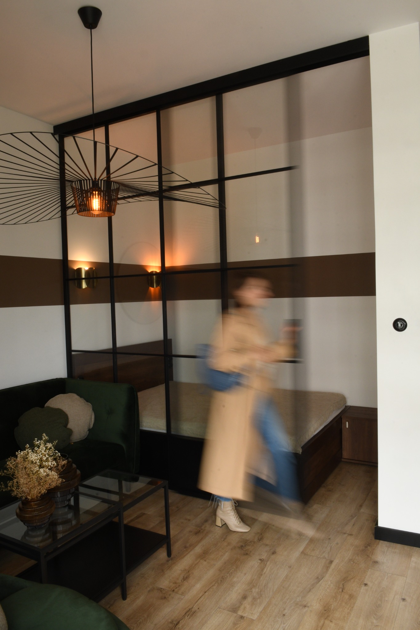 Nowoczesny salon z zieloną sofą, szklaną ścianką działową z czarnymi ramami, designerską lampą wiszącą i rozmytą sylwetką osoby w płaszczu.