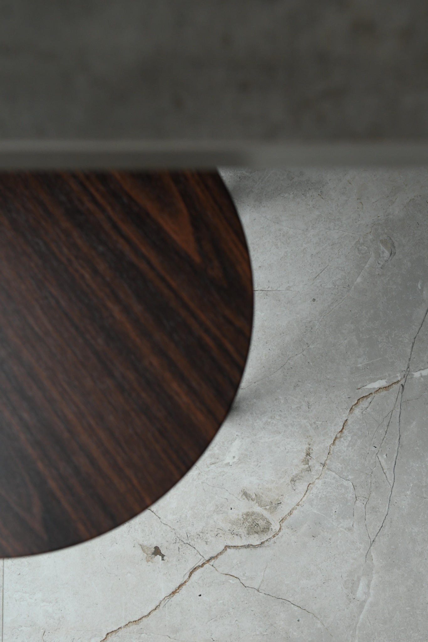 Fragment okrągłego stolika z ciemnego drewna na marmurowej podłodze w odcieniach szarości, widok z góry.