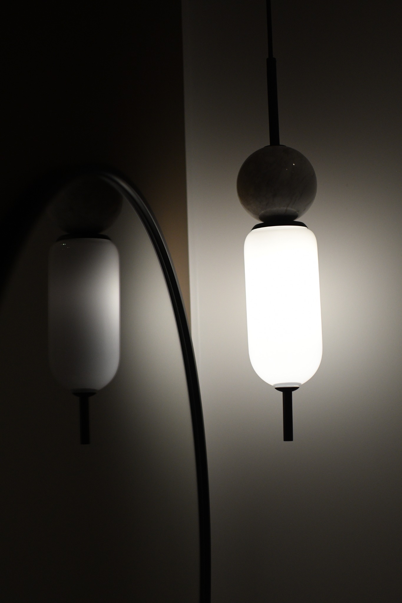 Nowoczesna lampa wisząca z marmurową kulą i mlecznym kloszem cylindrycznym, odbicie w łuku lustra, oświetlona ściana w tle.