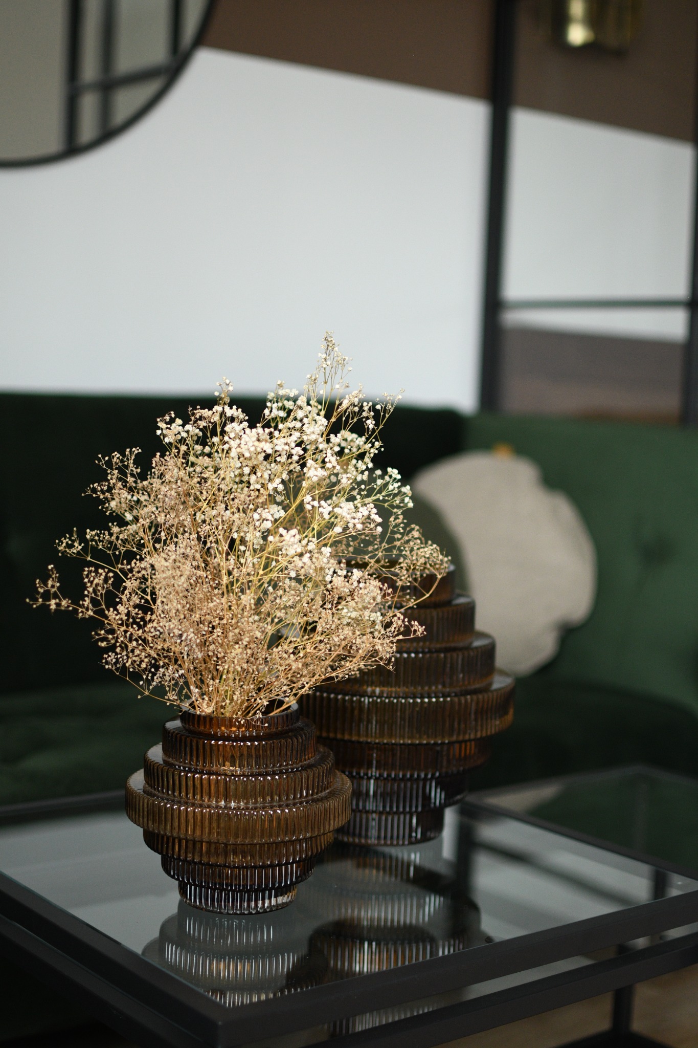 Dwa wazony z ciemnego szkła o żłobkowanej fakturze, jeden z suszonymi kwiatami gipsówki, stojące na szklanym stoliku kawowym z metalową ramą, w tle zielona sofa i okrągłe lustro.