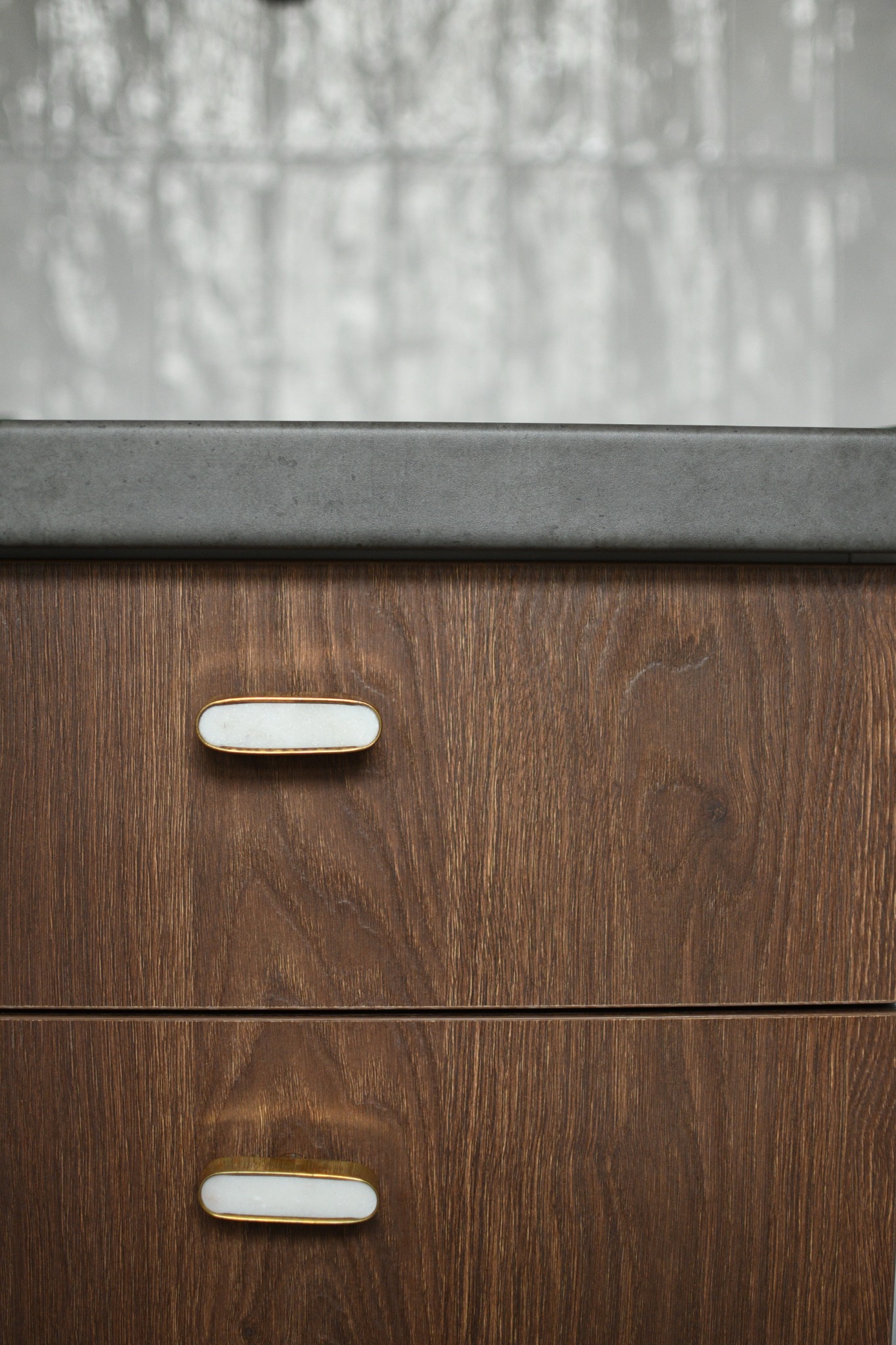 Szczegółowe ujęcie ciemnobrązowej drewnianej szafki z dwoma szufladami i marmurowymi uchwytami w złotej oprawie, na blacie z szarego kamienia, z rozmytym tłem w odcieniach szarości.