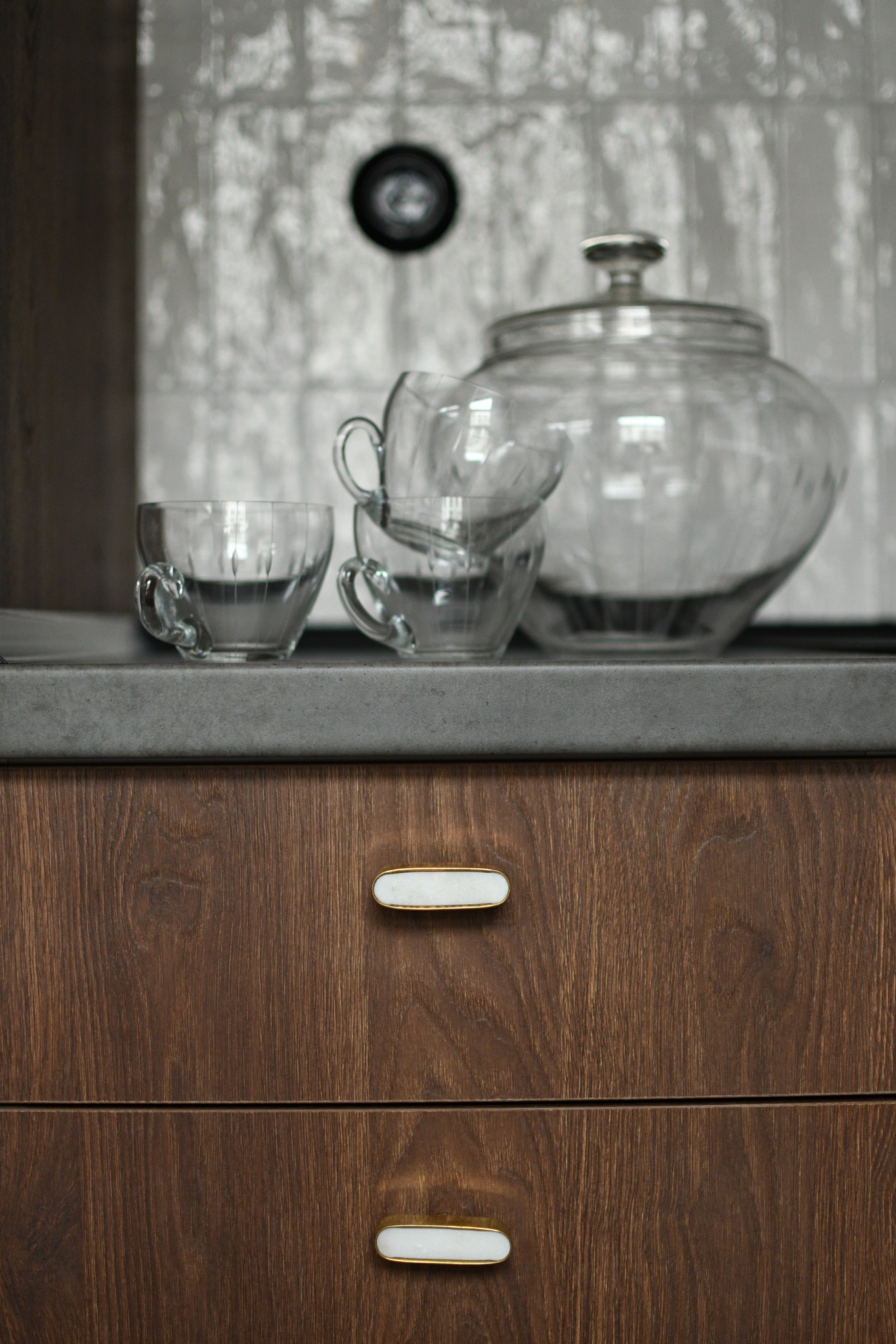 Szklane filiżanki i duży słój na blacie z ciemnego kamienia nad drewnianymi szufladami z ozdobnymi uchwytami w kuchni.