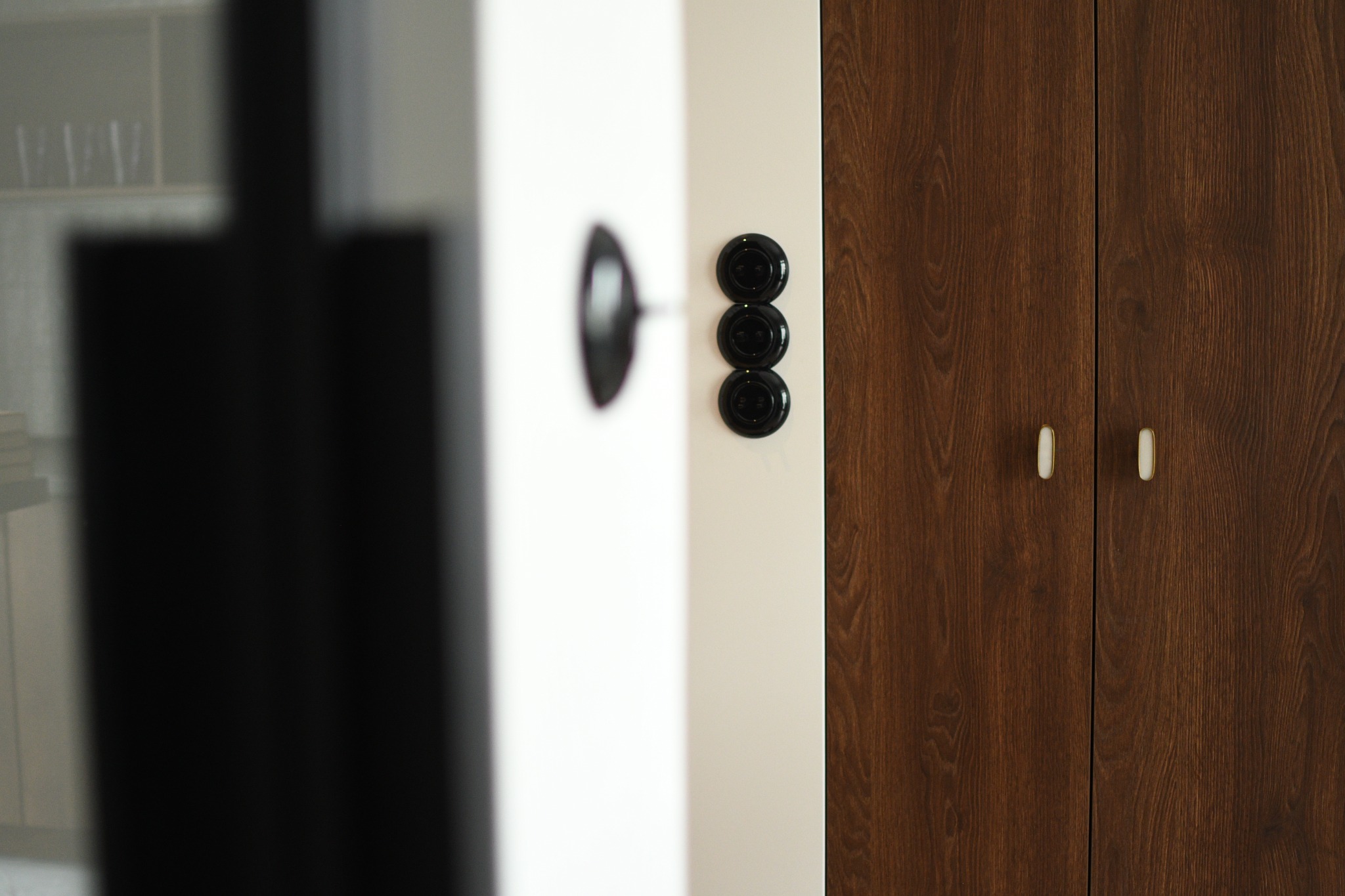 Fragment wnętrza: drewniana szafa z jasnymi uchwytami, biała ściana z potrójnym czarnym włącznikiem światła, widok z perspektywy otwartego pomieszczenia.