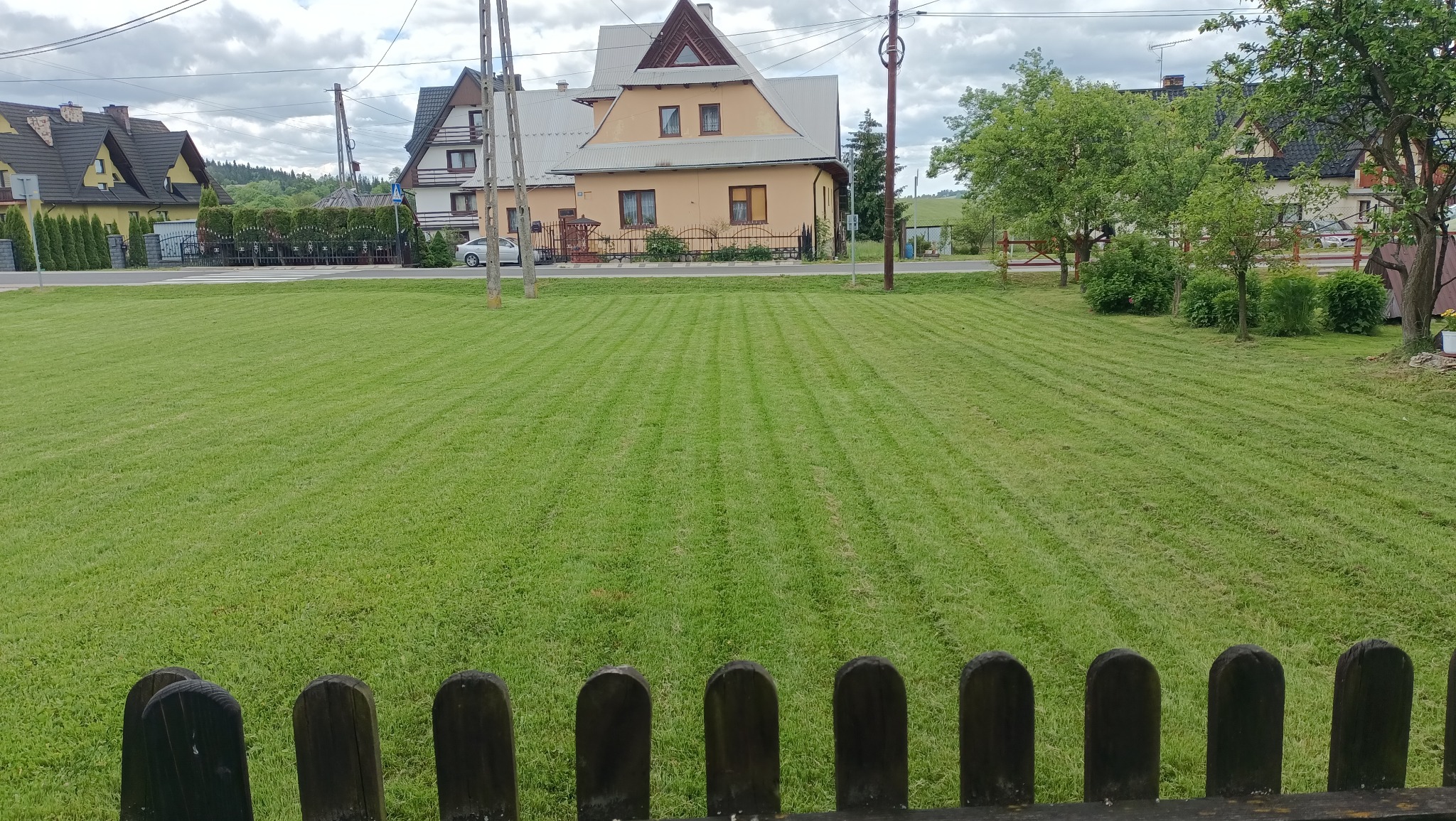 Równo skoszony trawnik z widocznymi pasami, w tle domy i słupy energetyczne, ujęcie znad drewnianego płotu.