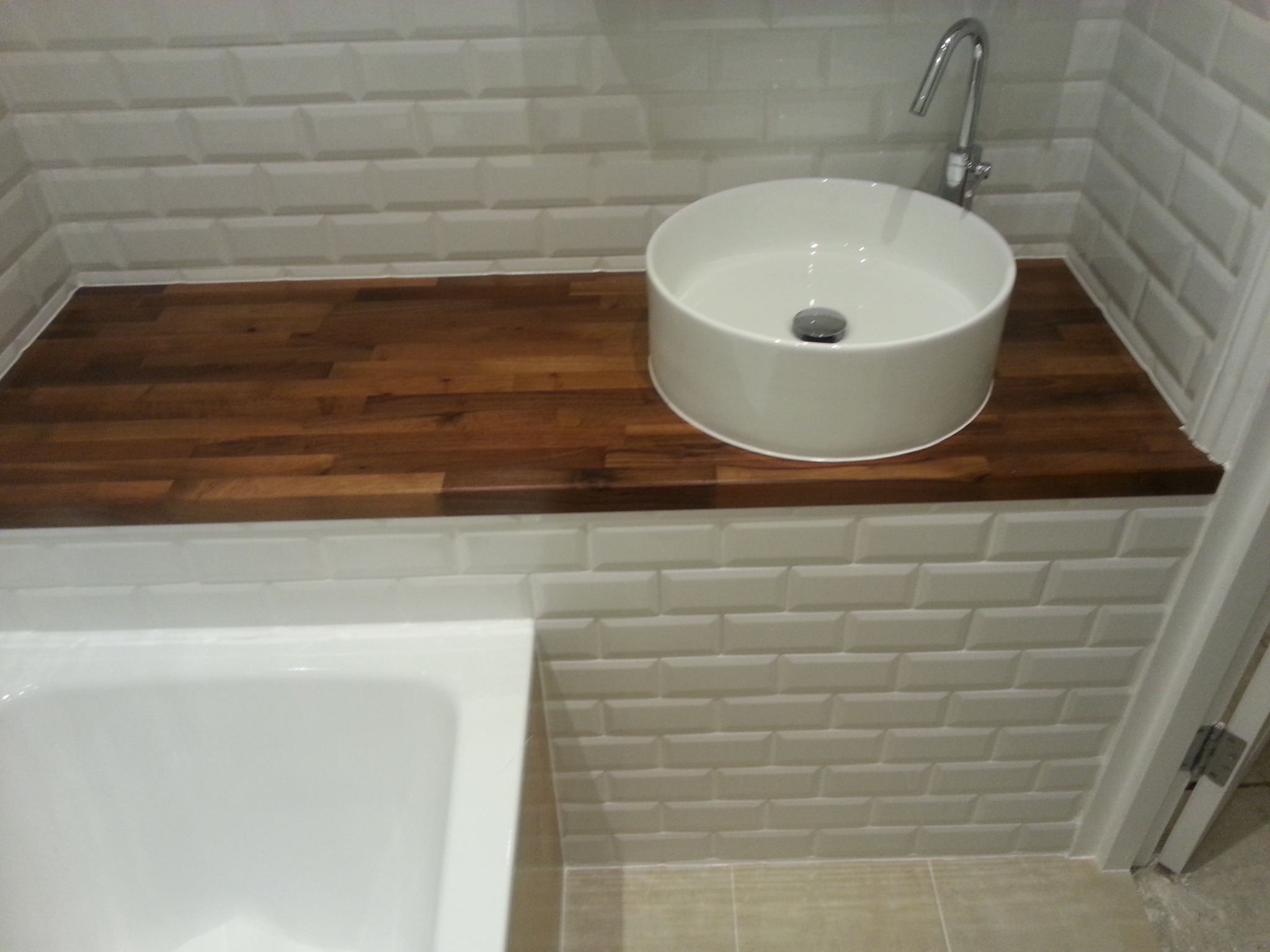 Biała, okrągła umywalka nablatowa z chromowaną baterią na blacie z ciemnego drewna, otoczona białymi, prostokątnymi płytkami imitującymi cegłę, z fragmentem wanny po lewej stronie.