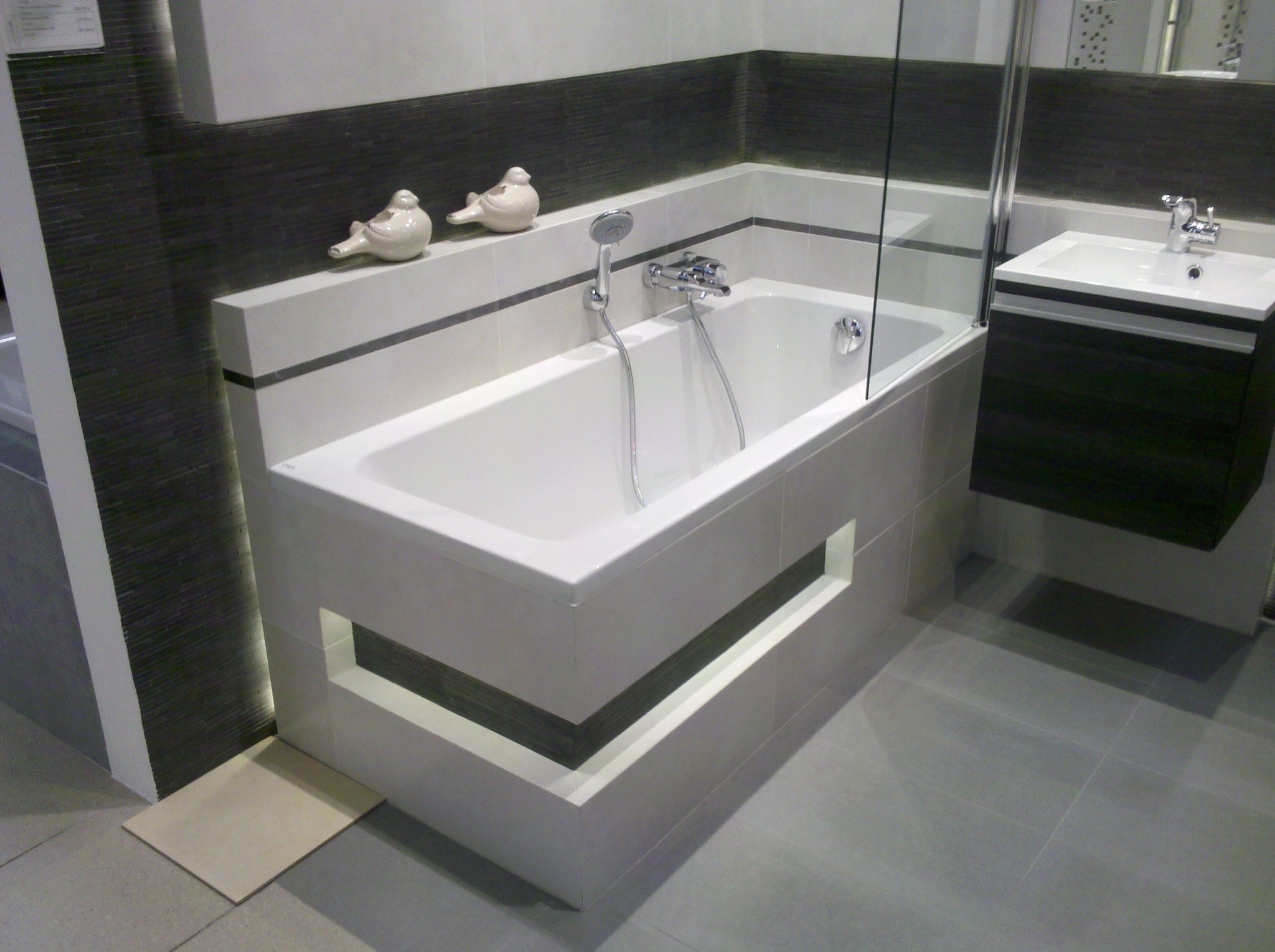 Wykończona łazienka z białą wanną obudowaną płytkami z dekoracyjnymi otworami, szara ściana wyłożona płytkami imitującymi drewno, dwie ceramiczne figurki ptaków i umywalka z szafką.