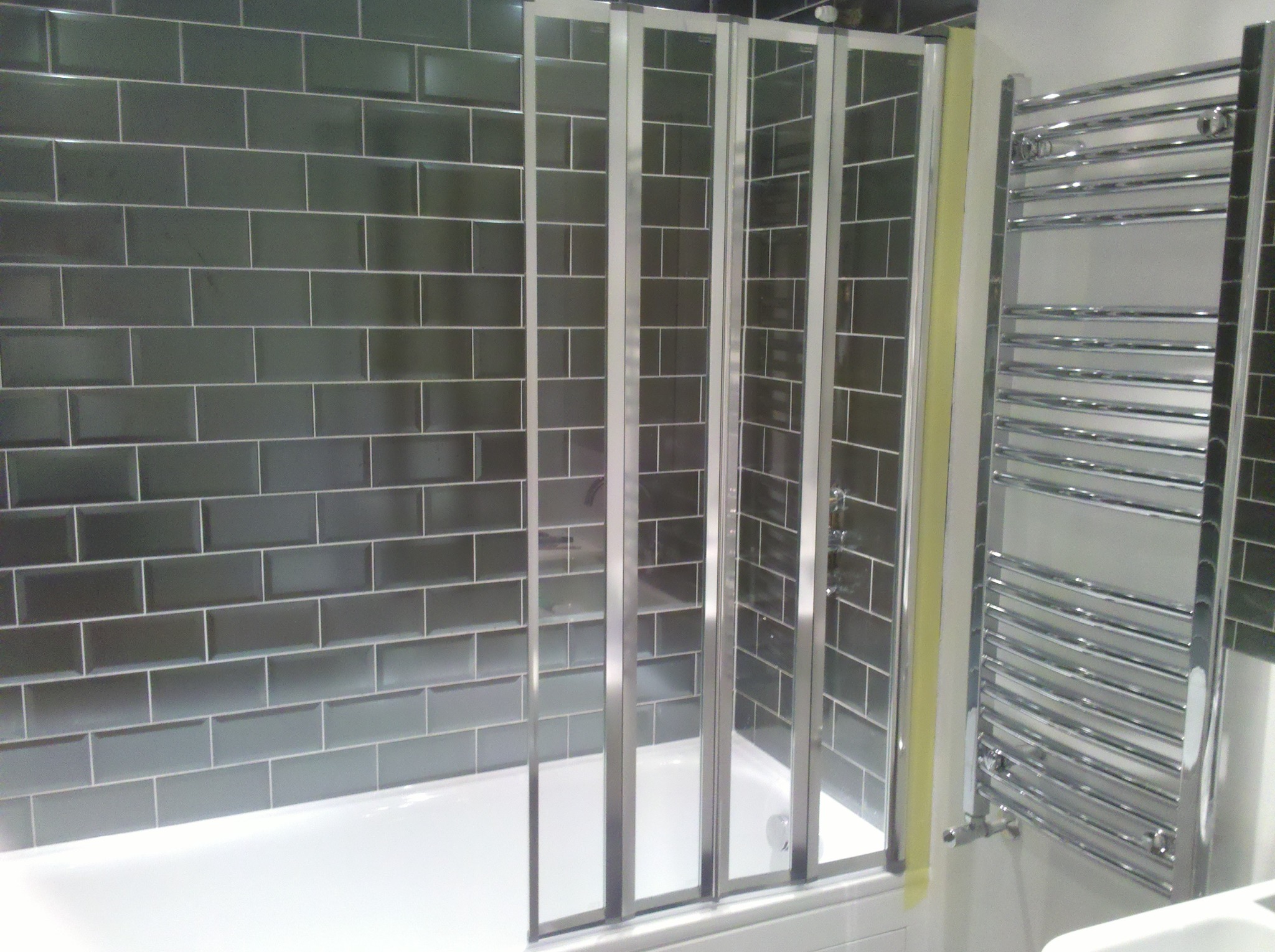 Wykończona łazienka z szarymi, prostokątnymi płytkami ułożonymi w cegiełkę, białą wanną z parawanem nawannowym i chromowanym grzejnikiem drabinkowym.